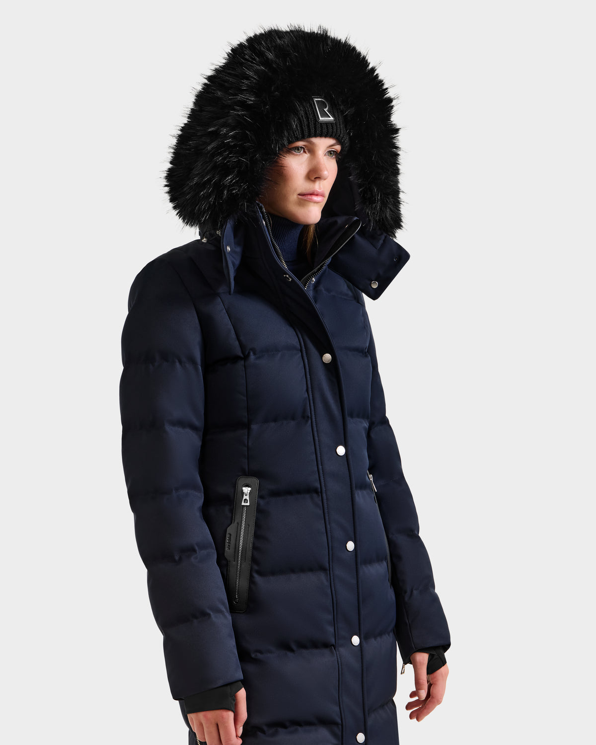 A woman in the Rudsak KEHLANI H - 8125643 Navy down parka with a fur-lined hood and black beanie stands against a white background. The coat features visible buttons, zippered pockets, and RDS-certified down for warmth.