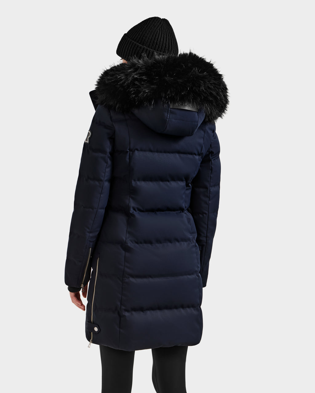 A person seen from the back wears the Rudsak KEHLANI H - 8125643 Navy women’s down parka with a black faux fur hood and a black knit beanie, standing against a plain light background—ideal for cold winter days.