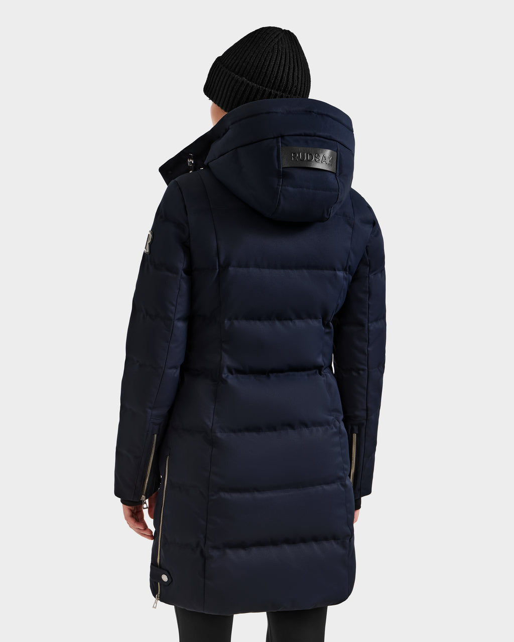 A person wearing the Rudsak KEHLANI H - 8125643 Navy women's down parka with a hood and a black knit beanie stands with their back to the camera, set against a plain light background.