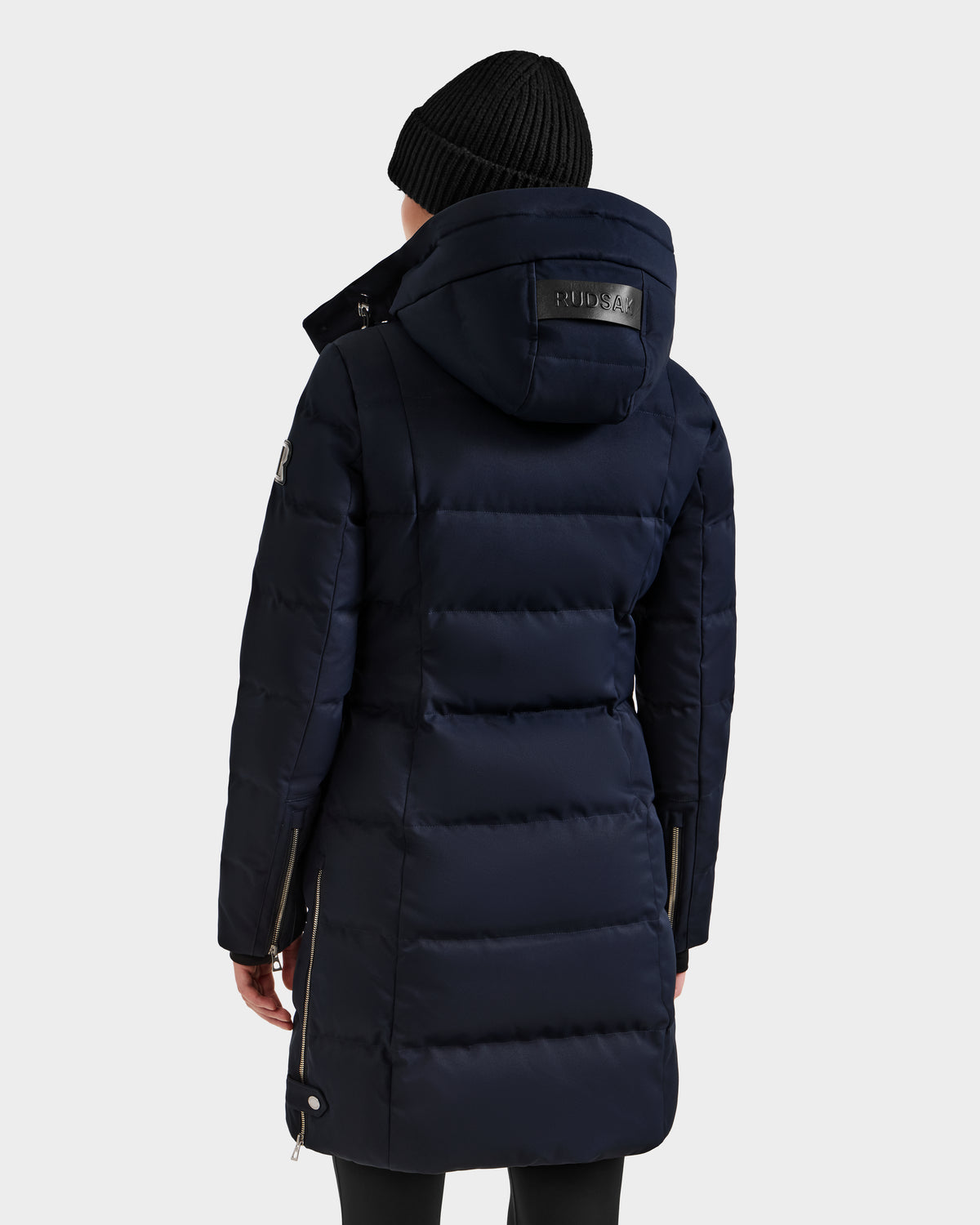 A person wearing the Rudsak KEHLANI H - 8125643 Navy women's down parka with a hood and a black knit beanie stands with their back to the camera, set against a plain light background.