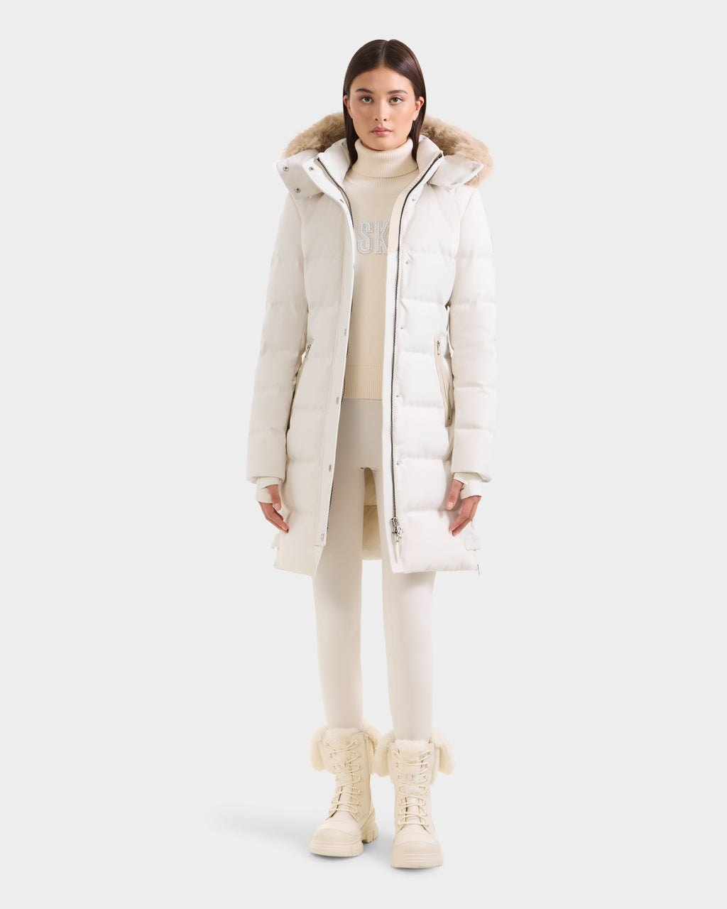 A woman stands facing forward wearing the Rudsak KEHLANI H - 8125643 Vanilla, a long white down parka with a fur-lined hood, over a cream sweater and leggings, paired with matching cream boots featuring fur trim, against a plain light background.