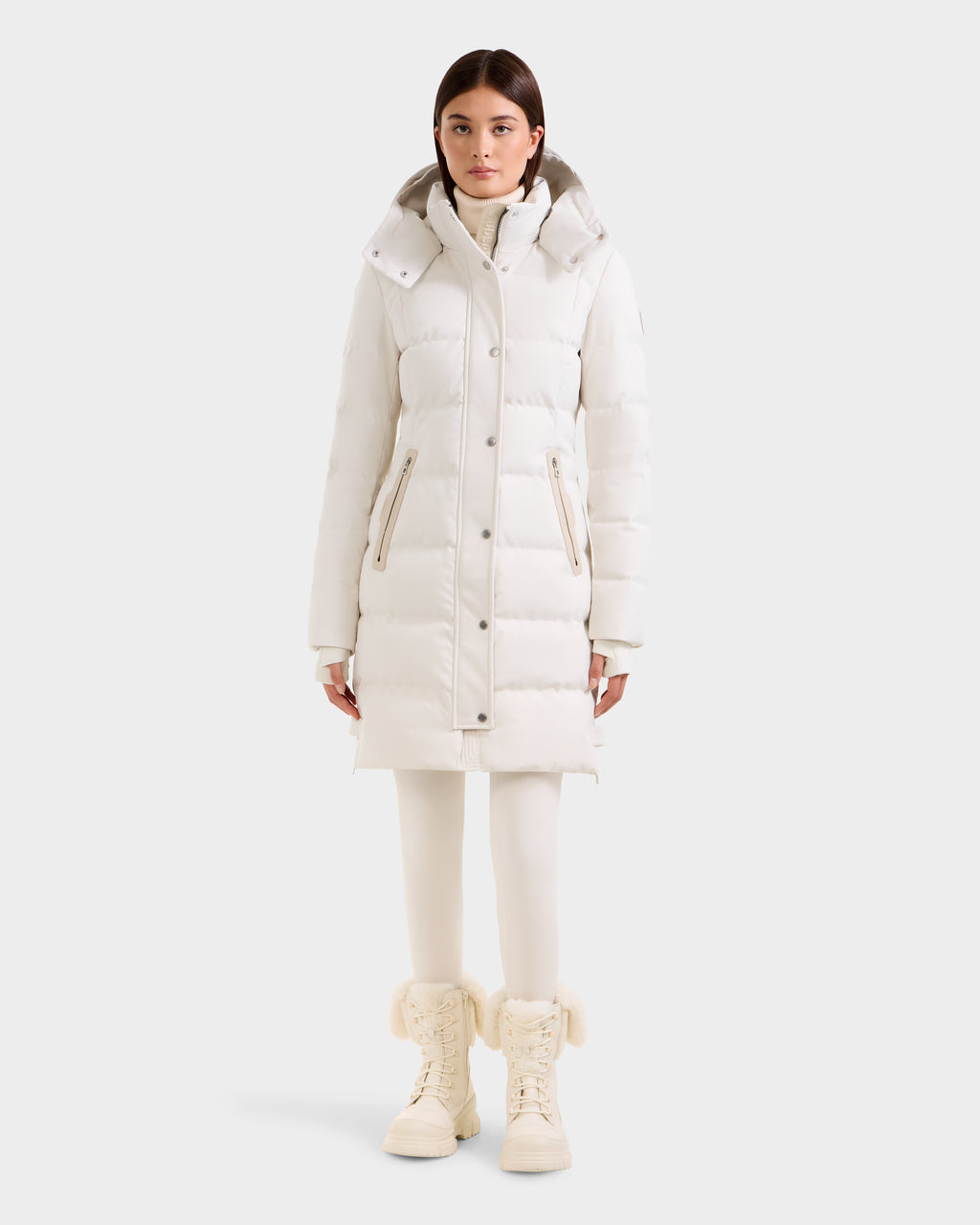A woman faces forward in a Rudsak KEHLANI H - 8125643 Vanilla knee-length down parka with hood, paired with white leggings and fur-trimmed white winter boots, set against a plain light background.