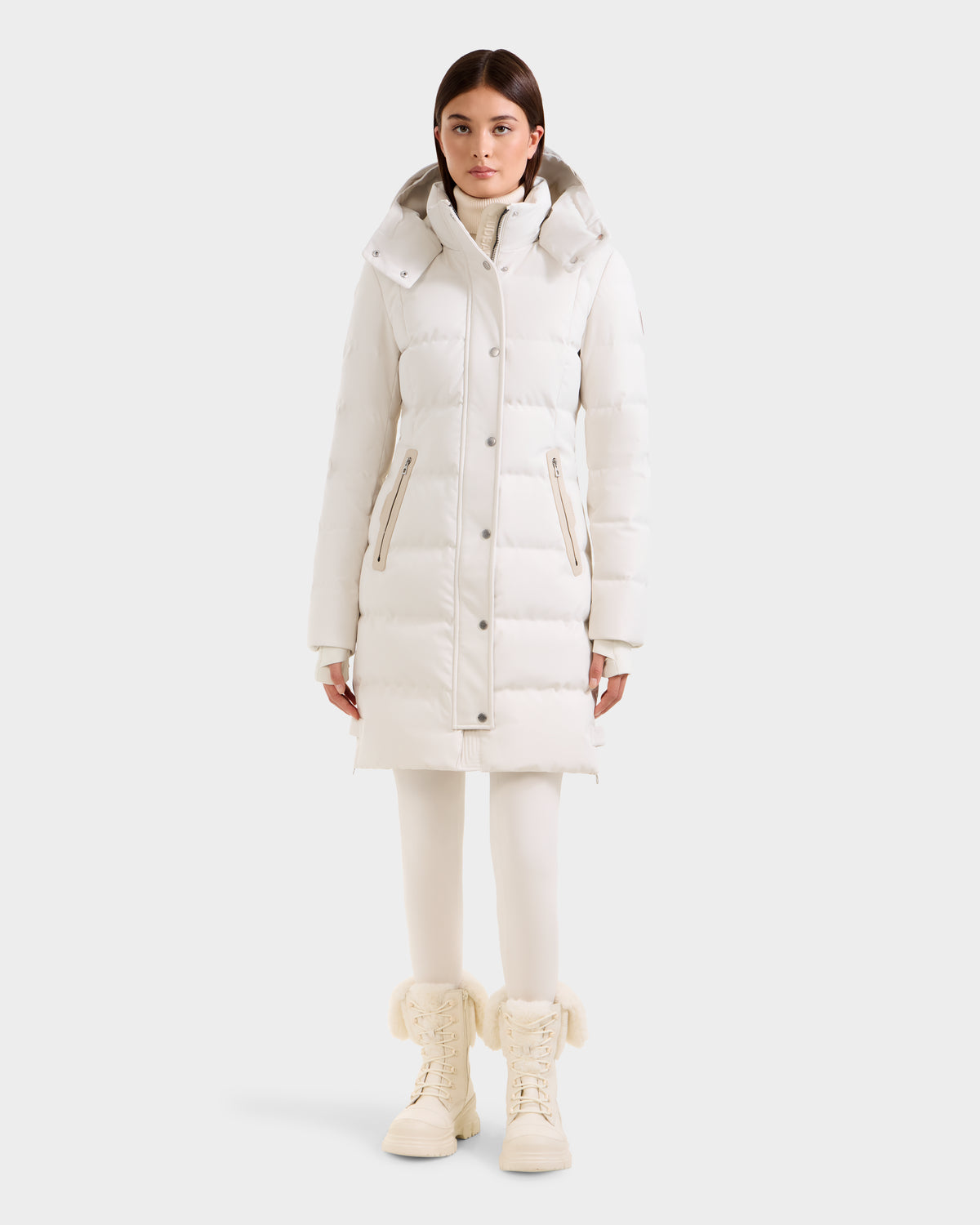 A woman faces forward in a Rudsak KEHLANI H - 8125643 Vanilla knee-length down parka with hood, paired with white leggings and fur-trimmed white winter boots, set against a plain light background.