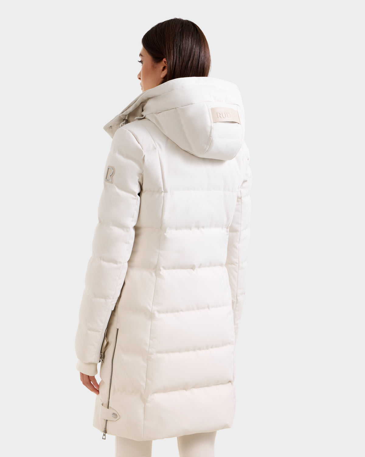 A person shown from the back and slightly turned to the side wears the Rudsak KEHLANI H - 8125643 Vanilla, a long white women’s down parka with a hood, against a plain light background.