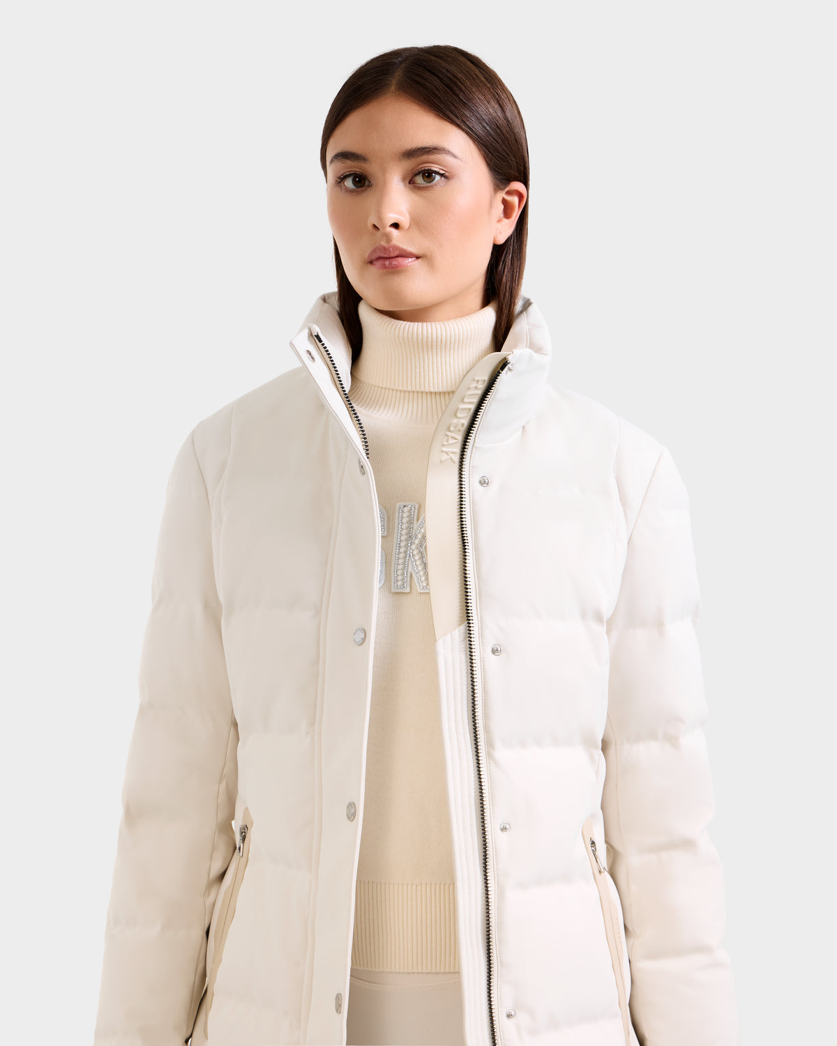 A woman with straight brown hair wears the Rudsak KEHLANI H - 8125643 Vanilla women’s down parka over a matching turtleneck sweater with silver lettering, standing against a plain light background.