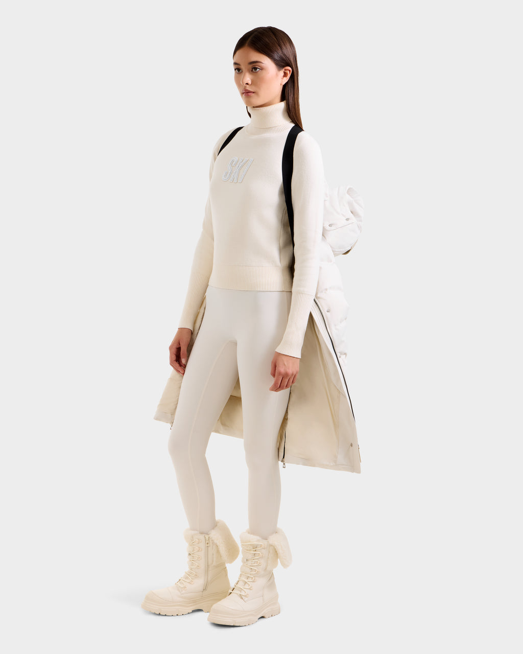 A woman poses against a plain background in a white turtleneck sweater with "SKI," white leggings, cream winter boots, and the Rudsak KEHLANI H - 8125643 Vanilla women's parka draped over her shoulders like a backpack.