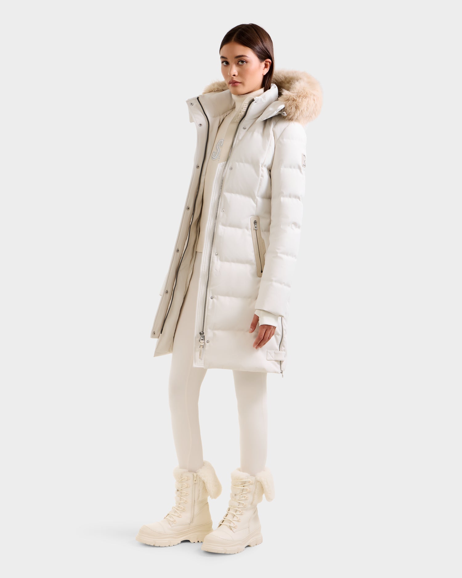 A woman wears the Rudsak KEHLANI H - 8125643 Vanilla RDS-certified down coat with a fur-trimmed hood, paired with a white turtleneck, leggings, and fur-lined boots against a plain light background.