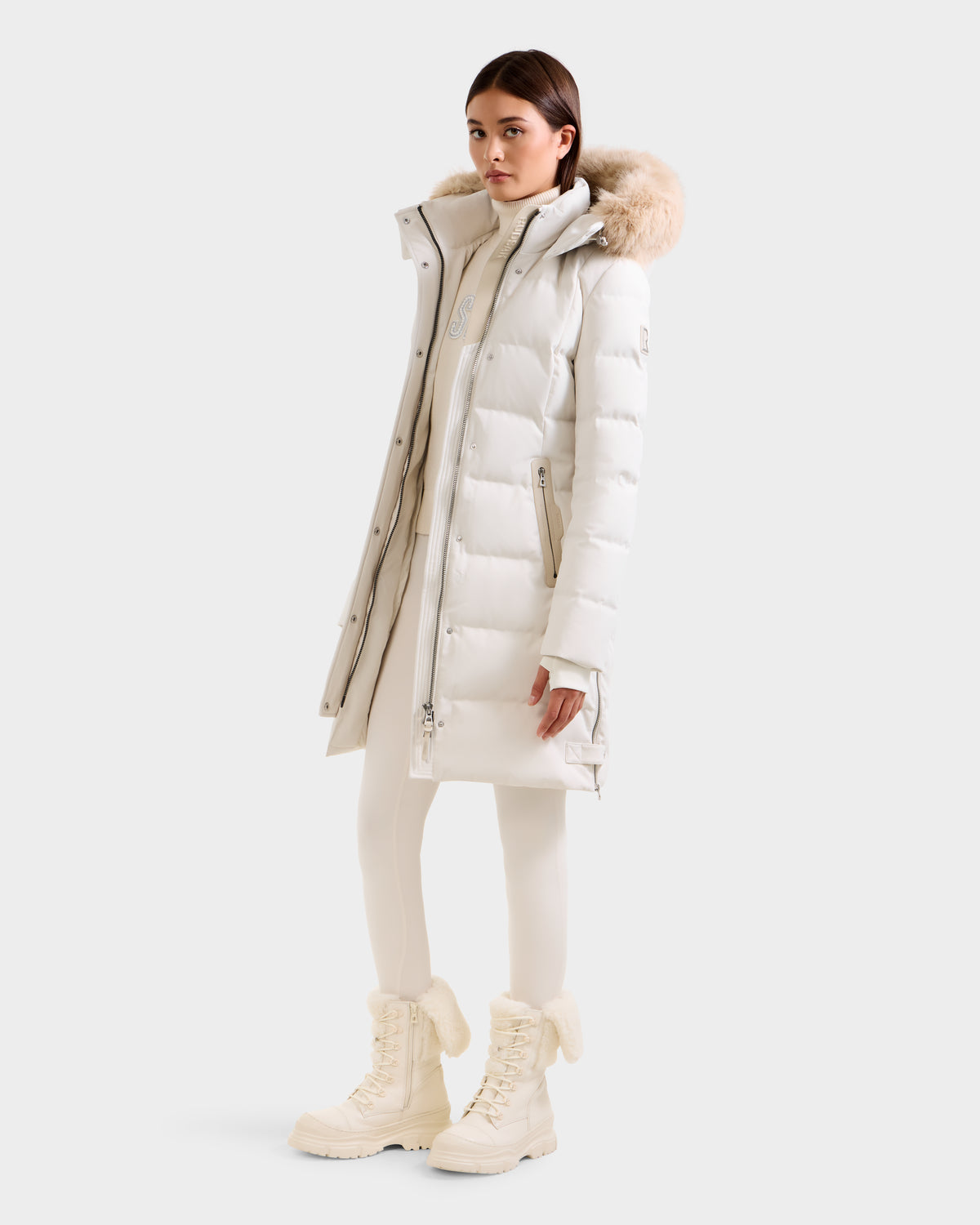 A woman wears the Rudsak KEHLANI H - 8125643 Vanilla RDS-certified down coat with a fur-trimmed hood, paired with a white turtleneck, leggings, and fur-lined boots against a plain light background.