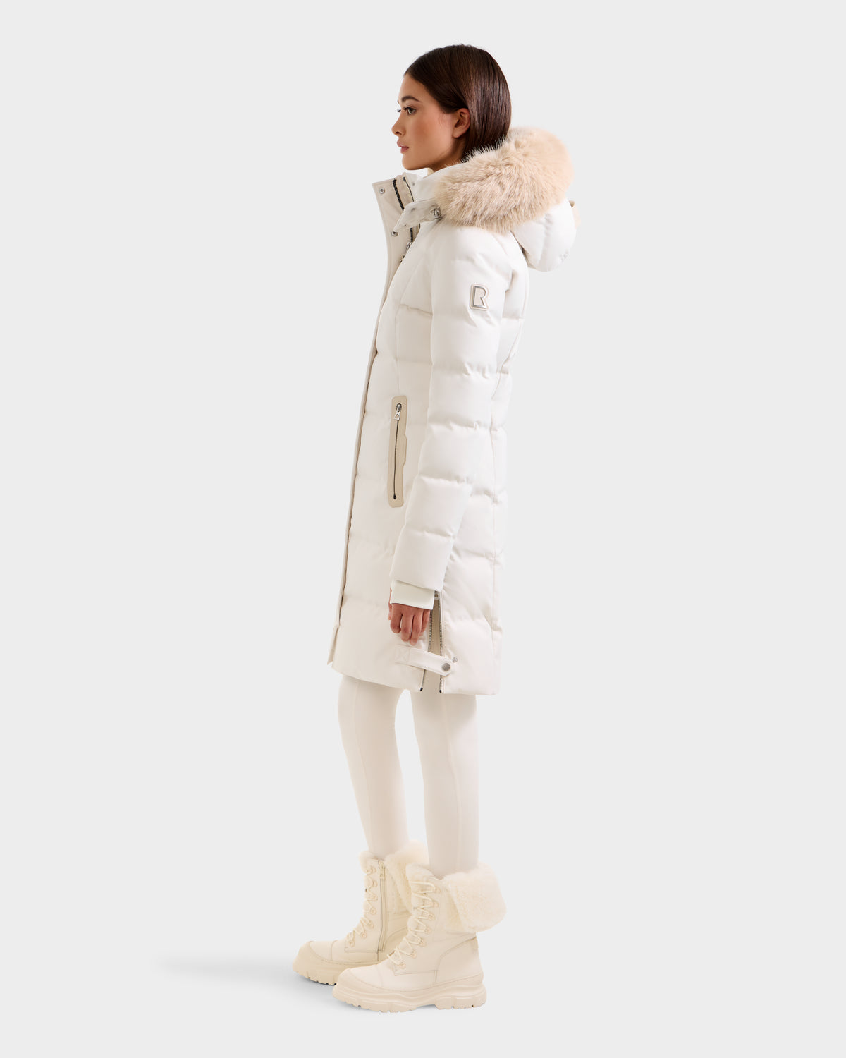 A woman wearing the Rudsak KEHLANI H - 8125643 Vanilla parka with fur-trimmed hood, white leggings, and fur-lined winter boots stands in profile against a plain light background.