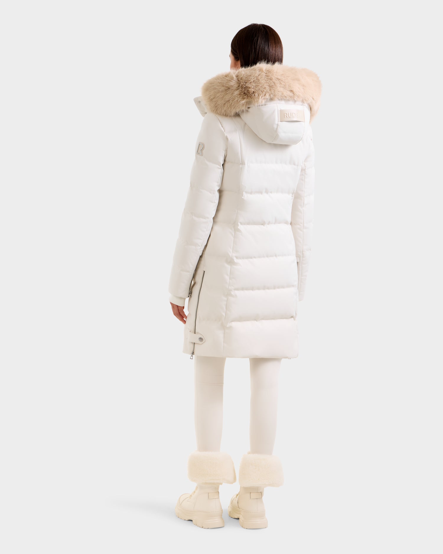 A person stands facing away, wearing the Rudsak KEHLANI H - 8125643 Vanilla women’s down parka with RDS-certified down and beige fur-trimmed hood, paired with white leggings and winter boots with fur cuffs against a plain light background.