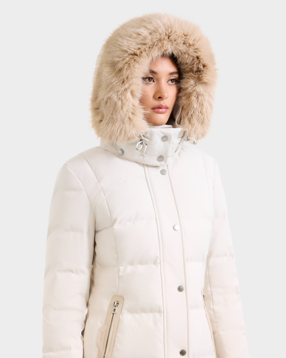 A person wears the Rudsak KEHLANI H - 8125643 Vanilla, a cream-colored quilted women's down parka with a large, fluffy fur-trimmed hood, looking slightly to the side against a plain light background.
