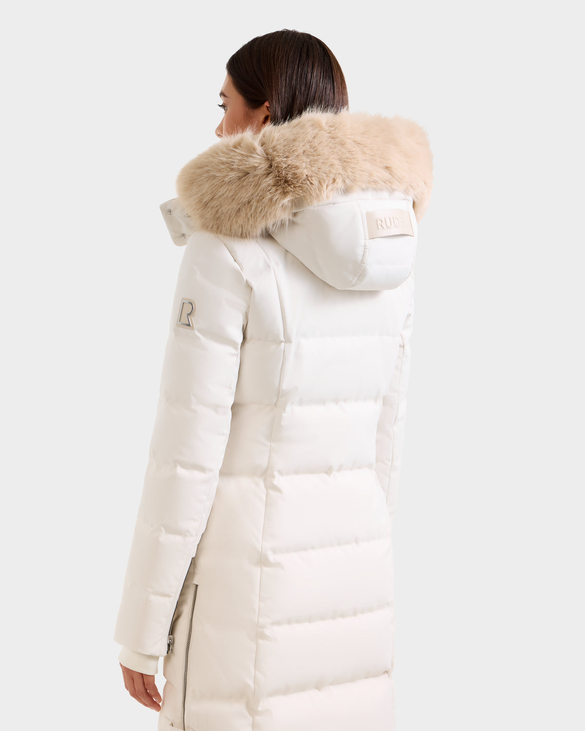 A woman with straight, dark brown hair faces away, wearing the Rudsak KEHLANI H - 8125643 Vanilla long white down parka, RDS-certified with a beige fur-trimmed hood and a visible R patch on the sleeve.