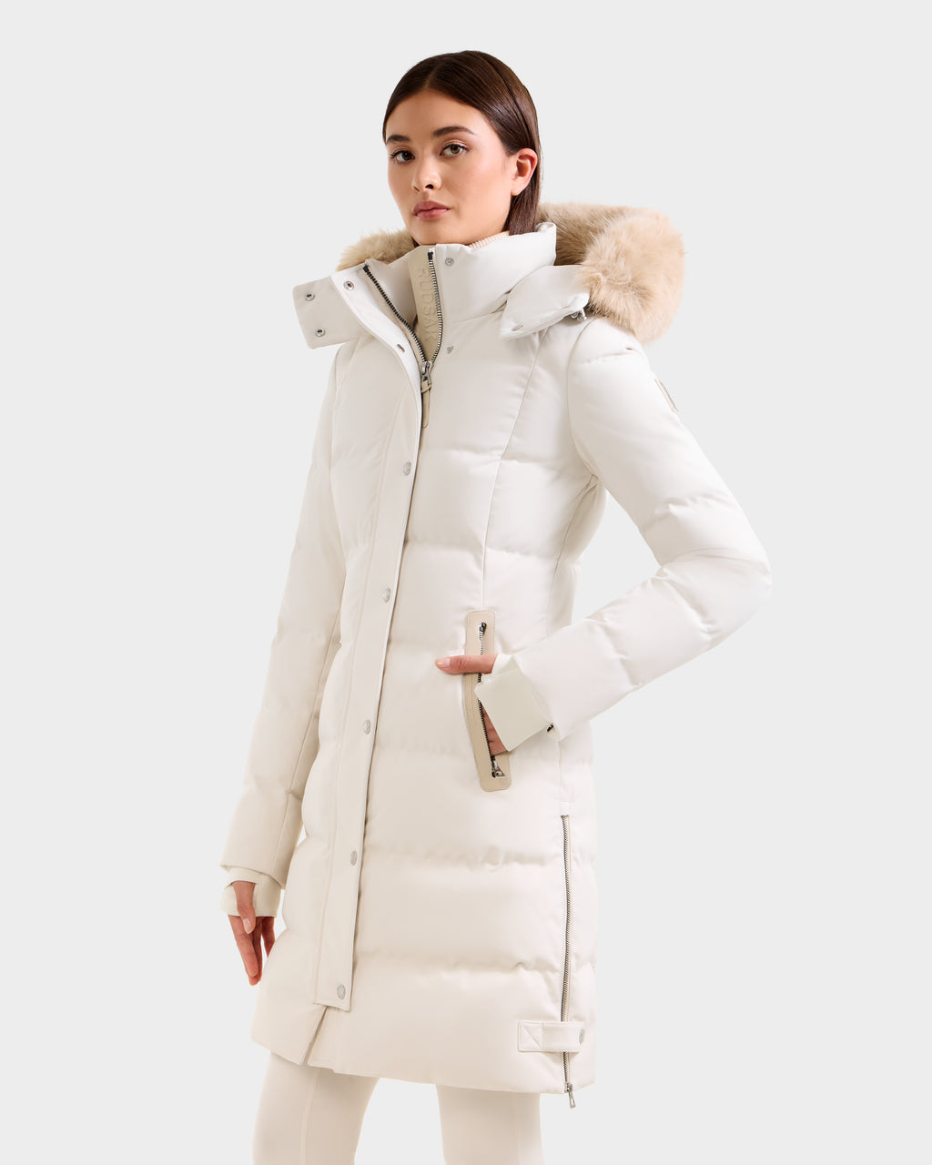 A woman with straight brown hair and a neutral expression wears the Rudsak KEHLANI H - 8125643 Vanilla, a long white down parka with a fur-trimmed hood, zipper, snap buttons, and zippered sleeve detail, standing against a plain light background.