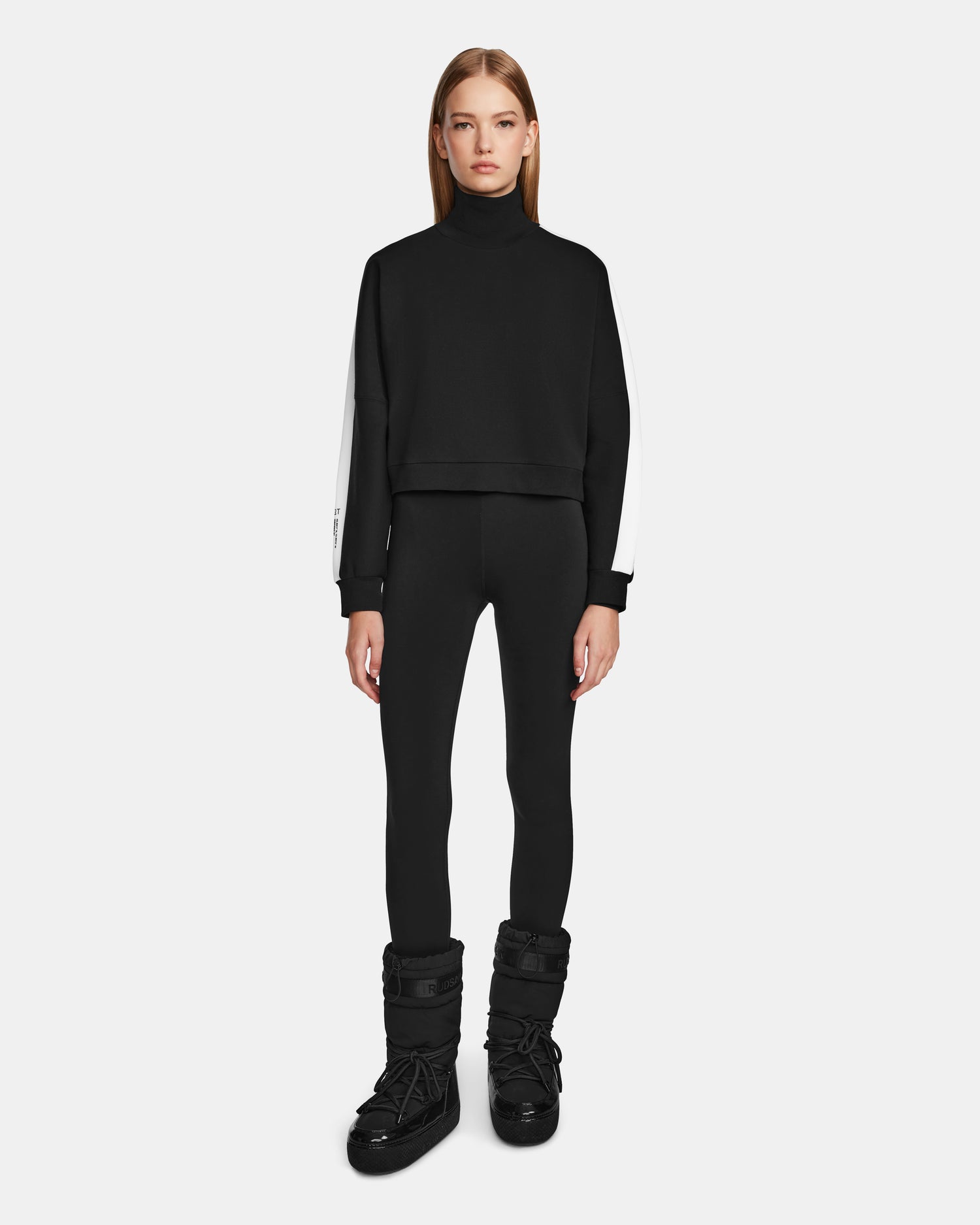 A woman stands facing forward against a light grey background, wearing the Rudsak KELLIE - 8524615 Black mock neck top with white sleeves, paired with black leggings and black winter boots.