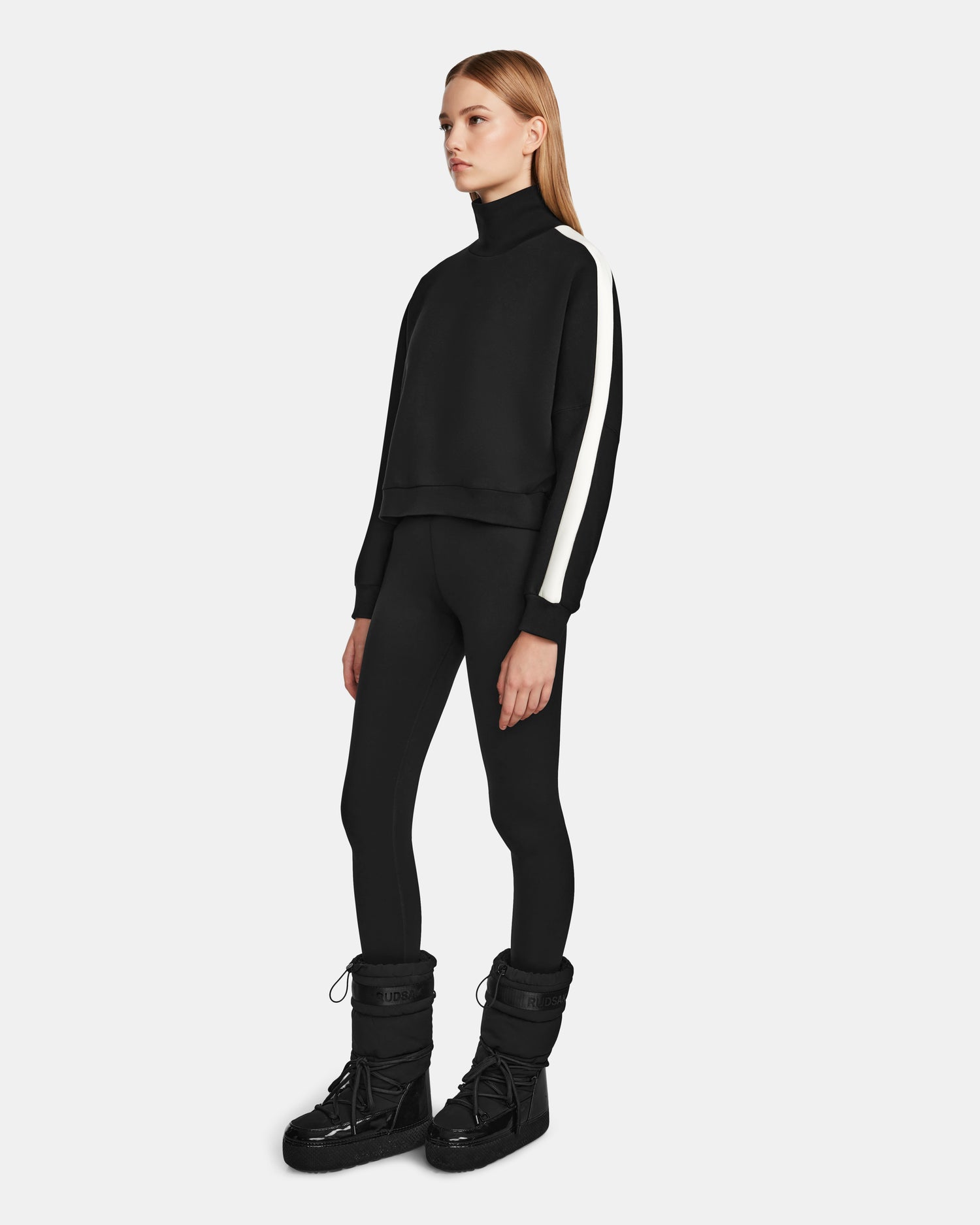 A woman faces slightly right, wearing the Rudsak KELLIE - 8524615 Black womens mock neck top with a white sleeve stripe, black leggings, and black winter boots against a plain white background.