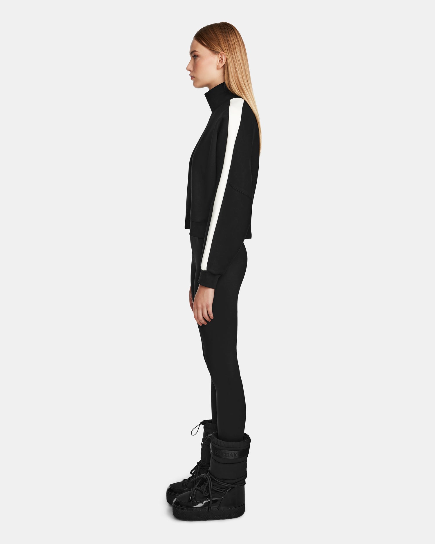 A woman stands in profile against a plain background, wearing the Rudsak KELLIE - 8524615 Black womens mock neck top with white sleeve stripes and black winter boots. Her straight hair is down as she looks straight ahead.