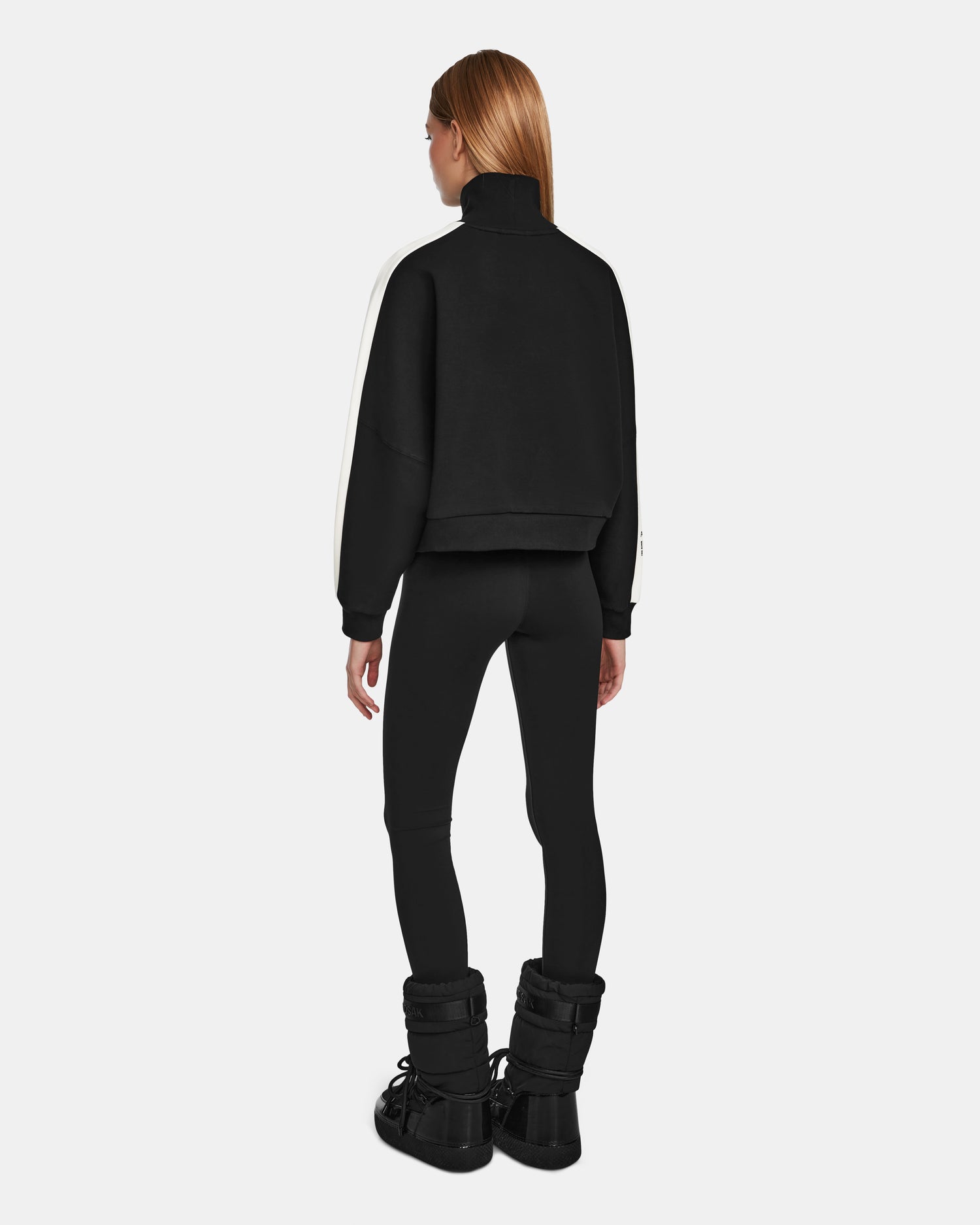 A woman with long, straight hair stands facing away, wearing the Rudsak KELLIE - 8524615 Black scuba fabric top with a white sleeve stripe, paired with black leggings and black winter boots against a plain white background.