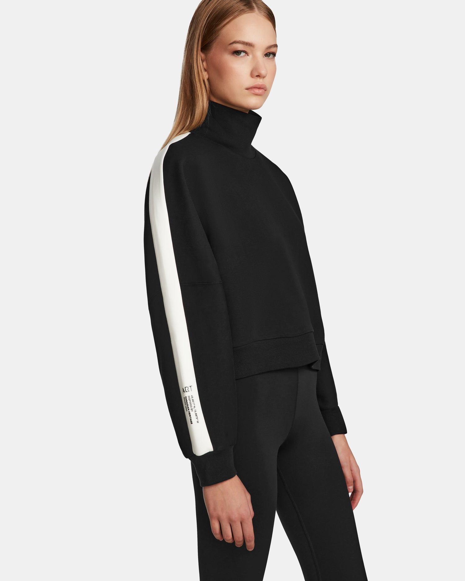 A woman wears the Rudsak KELLIE - 8524615 Black womens relaxed fit mock neck top with a white stripe down the sleeve, paired with black leggings, standing against a plain white background.