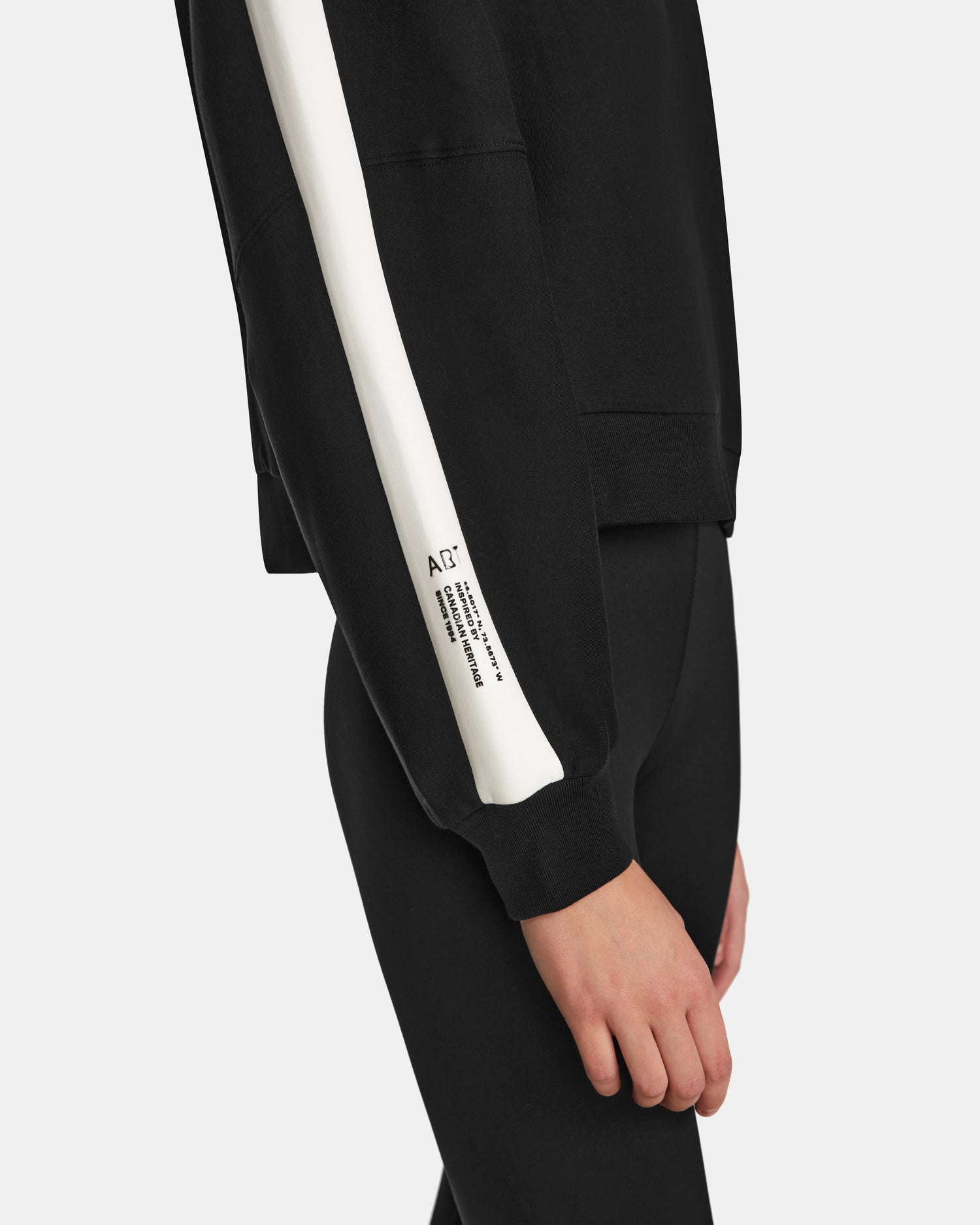 A person models the Rudsak KELLIE - 8524615 Black womens mock neck top—a black sweatshirt with a wide white sleeve stripe and small printed text near the wrist, styled with black pants against a white background.