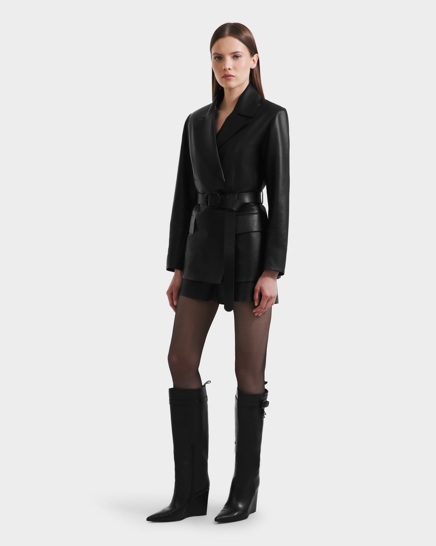 A woman stands facing forward against a light gray background, wearing the Rudsak KENDALL - 8125617 Black leather belted blazer, a black skirt, sheer black tights, and knee-high black boots.