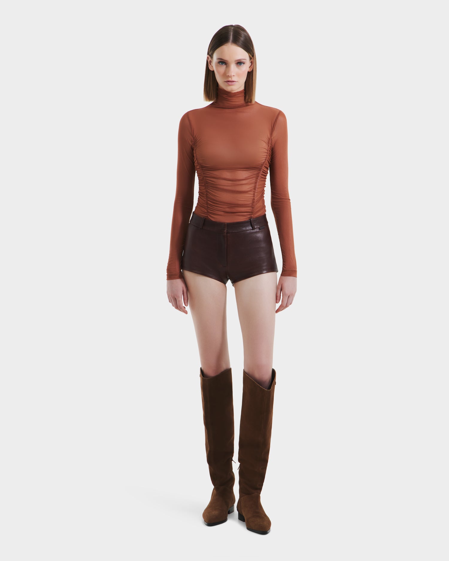 A woman faces forward against a plain white background, wearing Rudsaks KENNEDY - 8525610 Espresso tall boots, paired with a fitted, long-sleeve rust turtleneck and high-rise leather hot shorts.