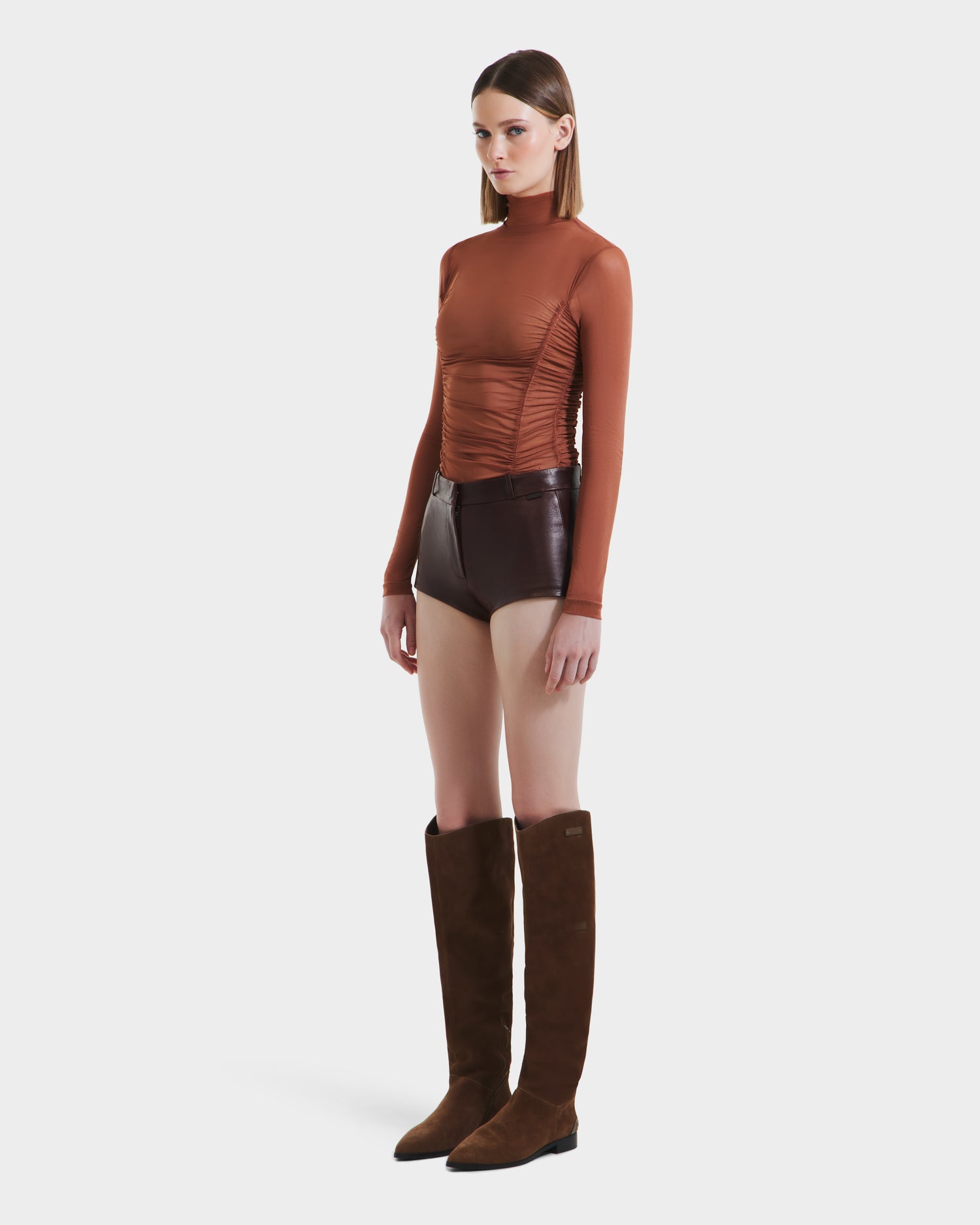 A woman with straight brown hair and a neutral expression wears the Rudsak KENNEDY - 8525610 Espresso: a fitted long-sleeve brown top, high-rise leather hot shorts, and tall brown suede boots, standing against a plain background.
