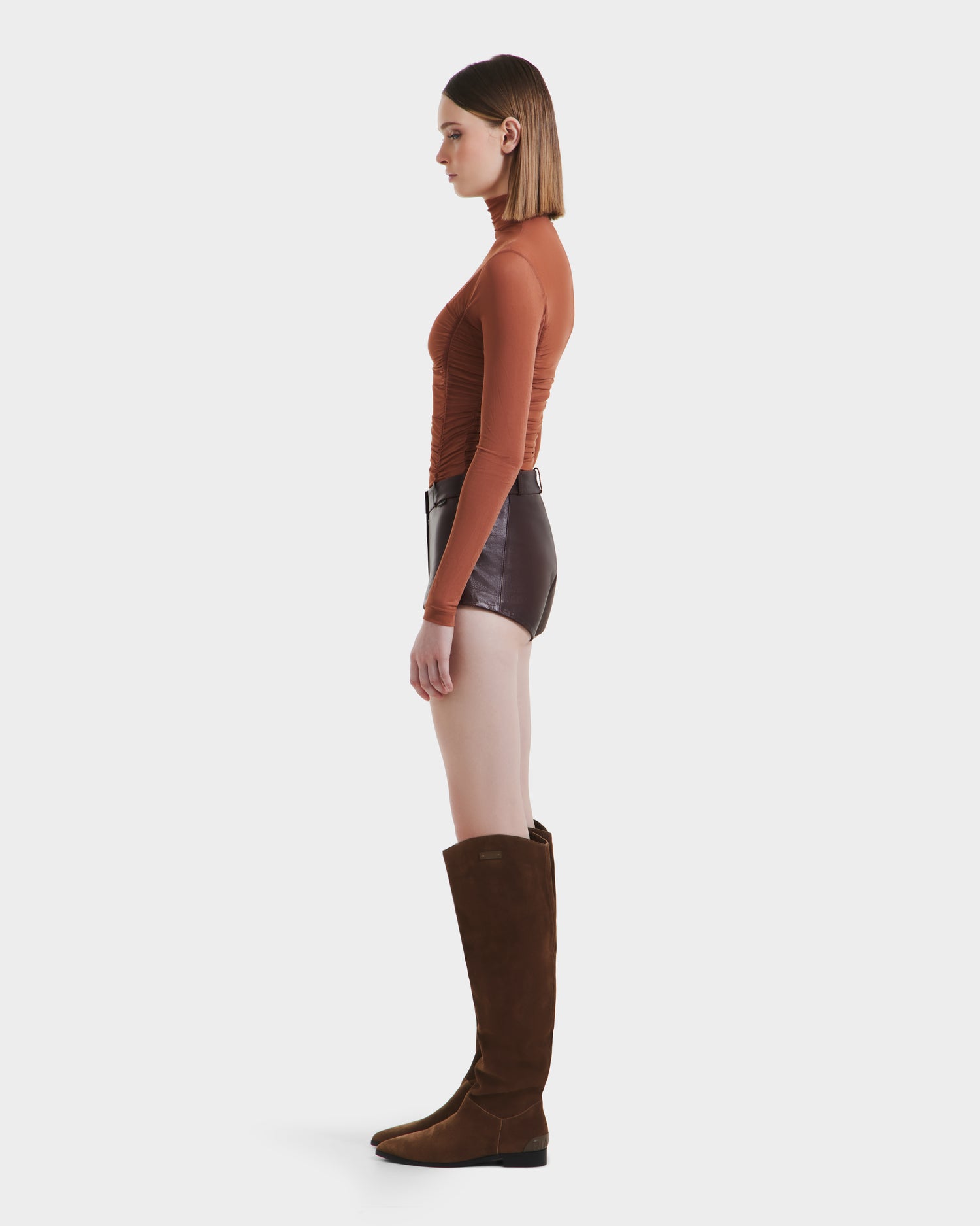 A woman stands in profile against a white background, wearing a fitted rust-colored long-sleeve top, high-rise women’s shorts, and KENNEDY - 8525610 Espresso knee-high boots by Rudsak.