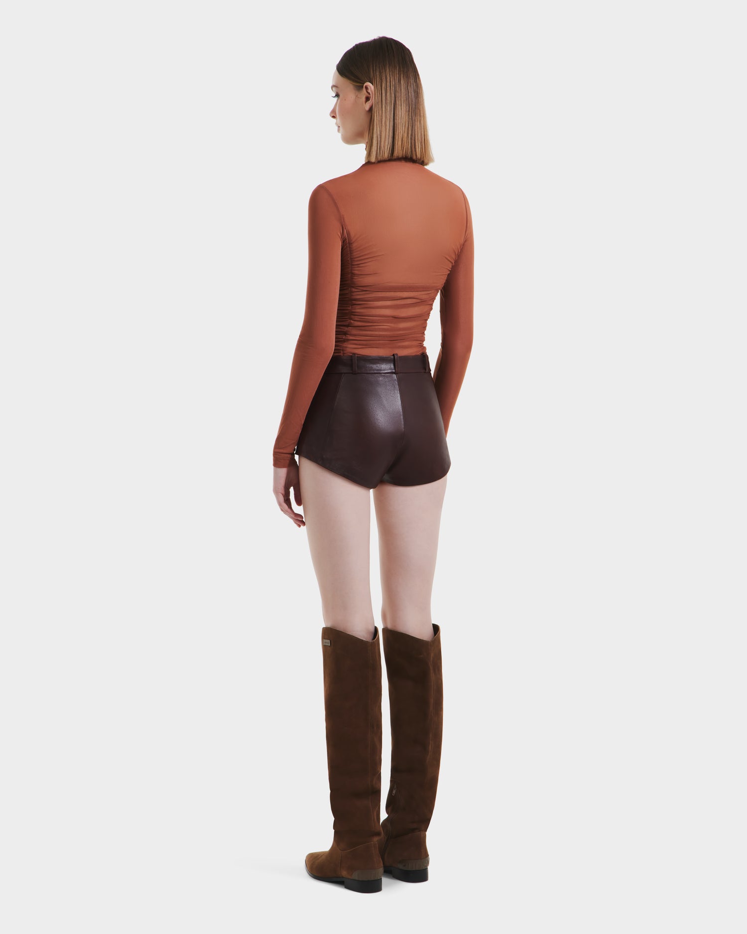 A woman faces away against a white background, wearing Rudsaks KENNEDY - 8525610 Espresso knee-high brown suede boots paired with a long-sleeve rust top and high-rise shorts.