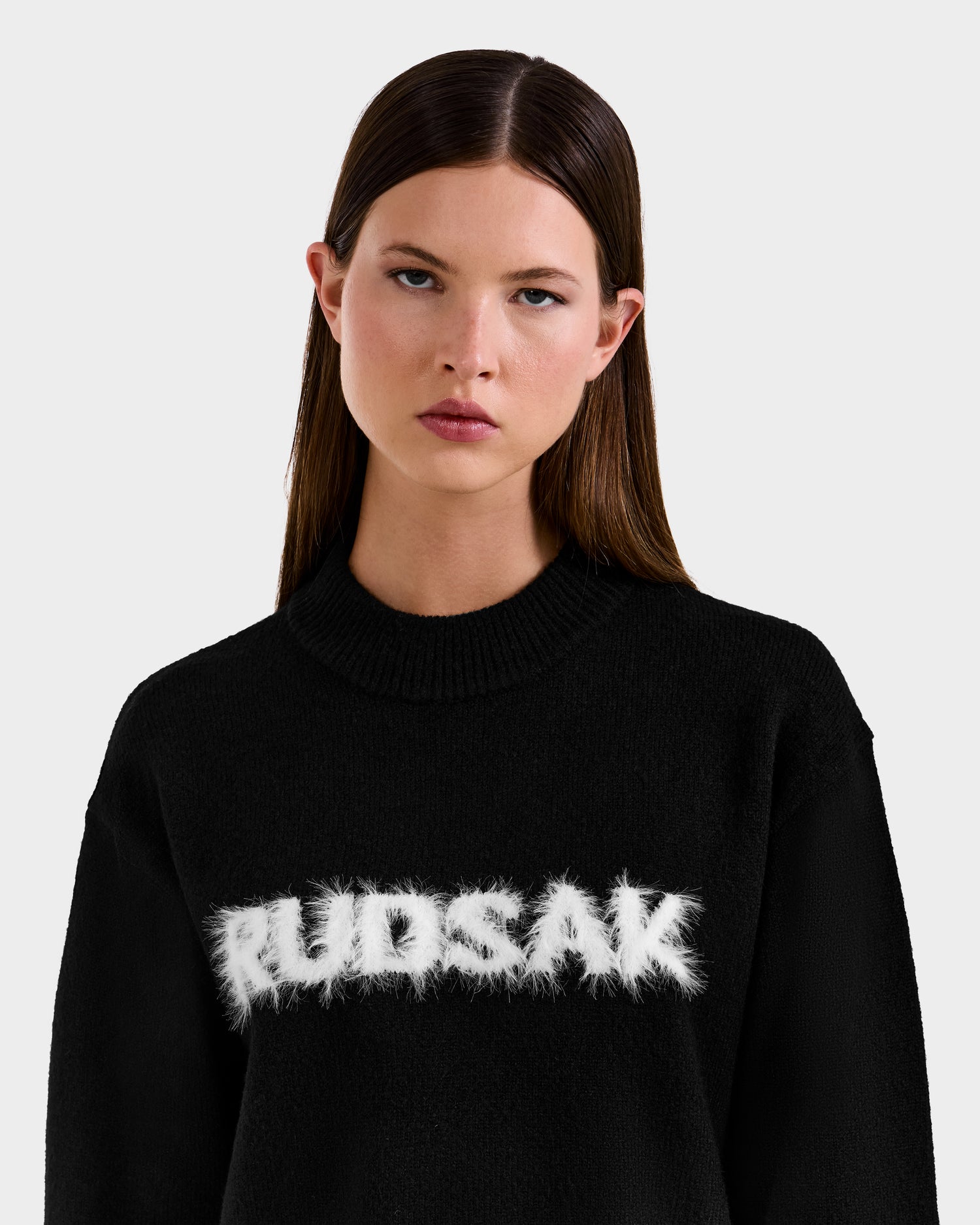 A woman with straight brown hair wears the Rudsak KINLEY - 9525555 Black W sweater, which features bold white logo letters across the chest. She stands against a light gray background, looking ahead with a neutral expression.