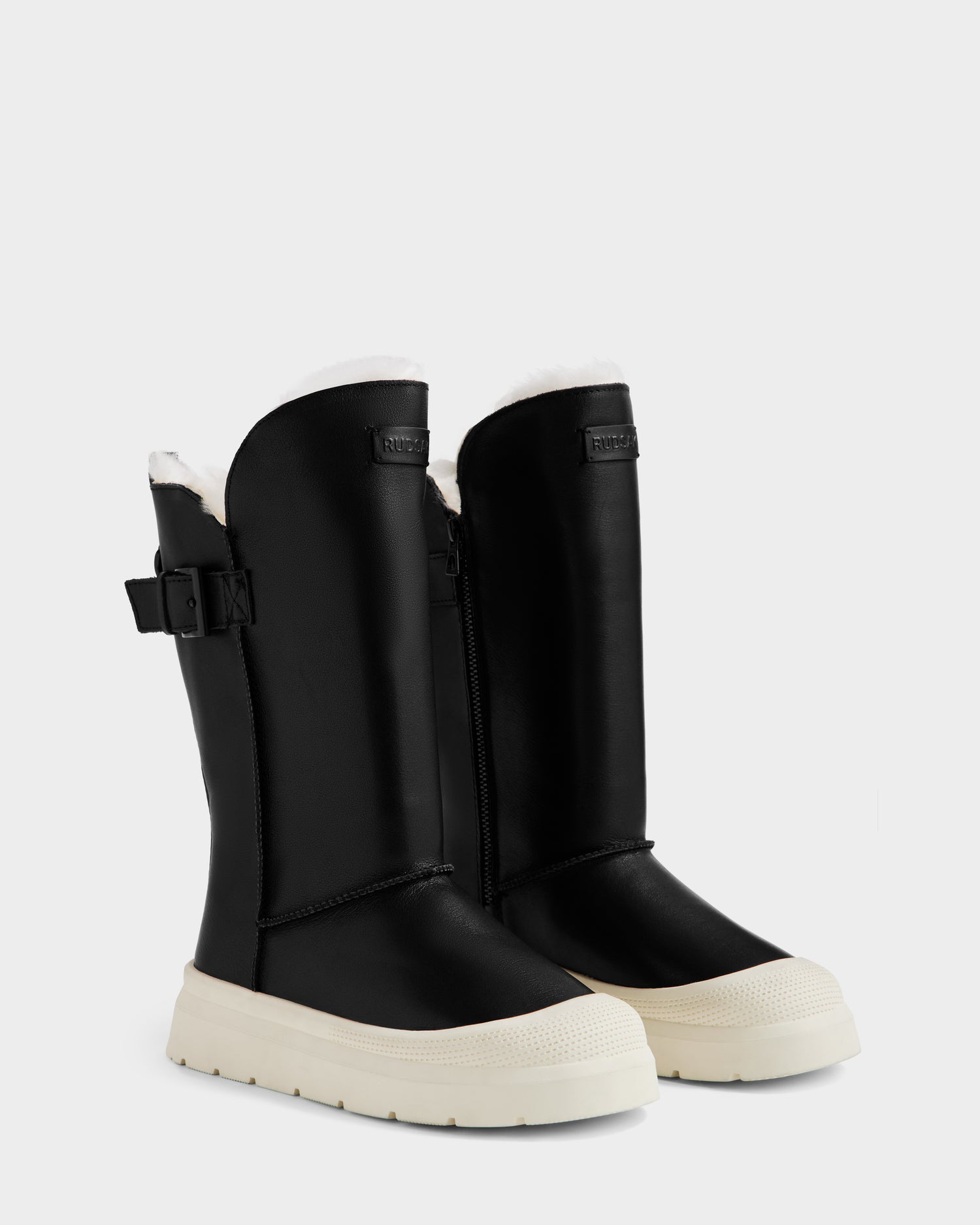 The Rudsak KLAIR L - L225519 Black women’s leather boots feature white soles, side zippers, adjustable side buckles, and cozy wool lining at the top—ideal for city traction. Shown on a plain light background.