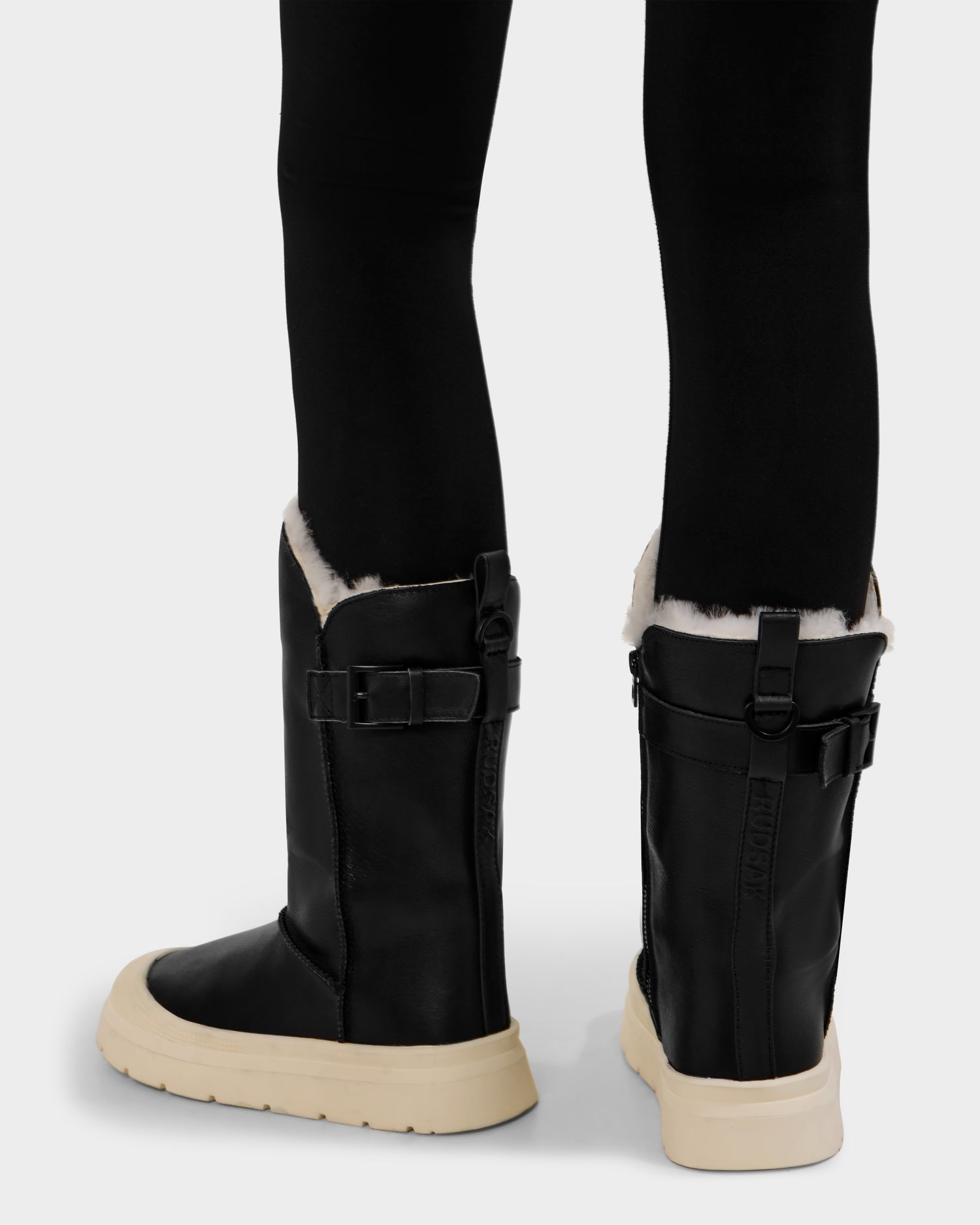 A person shown from the back wears Rudsak KLAIR L - L225519 Black mid-calf boots with side buckles, cream-colored soles, visible white fleece lining at the top, and black leggings.