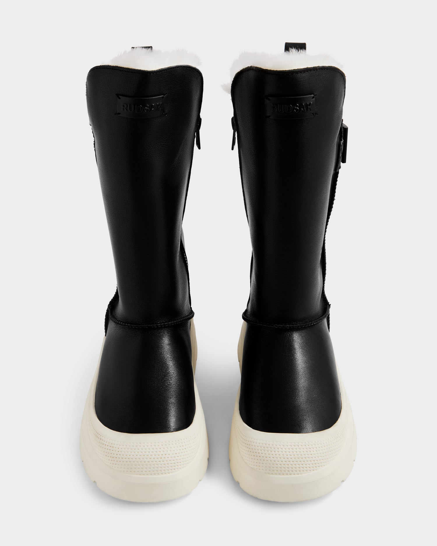 The Rudsak KLAIR L - L225519 Black women's leather boots feature black uppers, white soles, and a white fur lining for city-ready traction, shown from the back against a plain light gray background.
