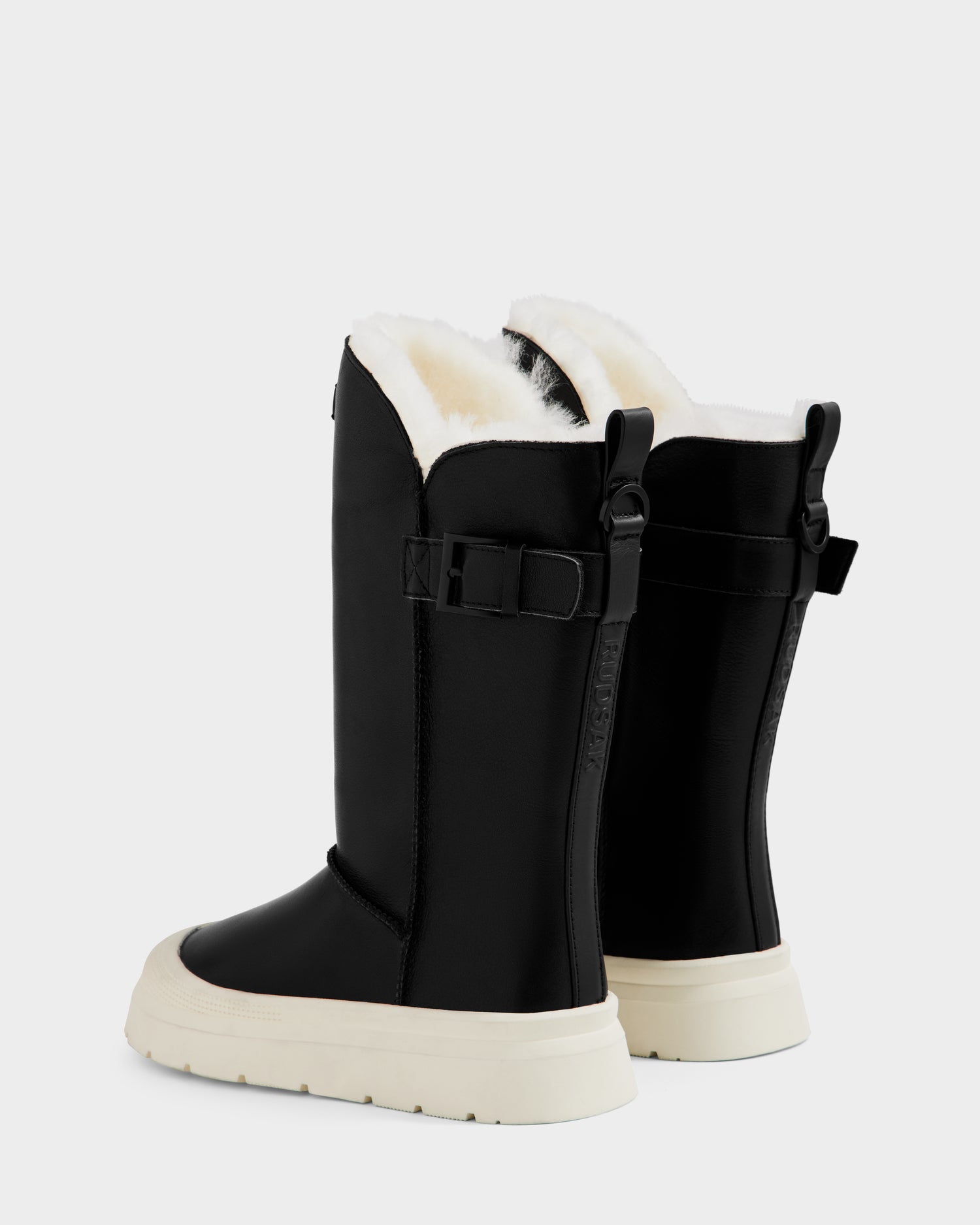 The Rudsak KLAIR L - L225519 Black women’s leather boots feature black wool lining, plush white interior, thick white soles, adjustable side buckles, and city-ready traction, displayed on a plain light background.