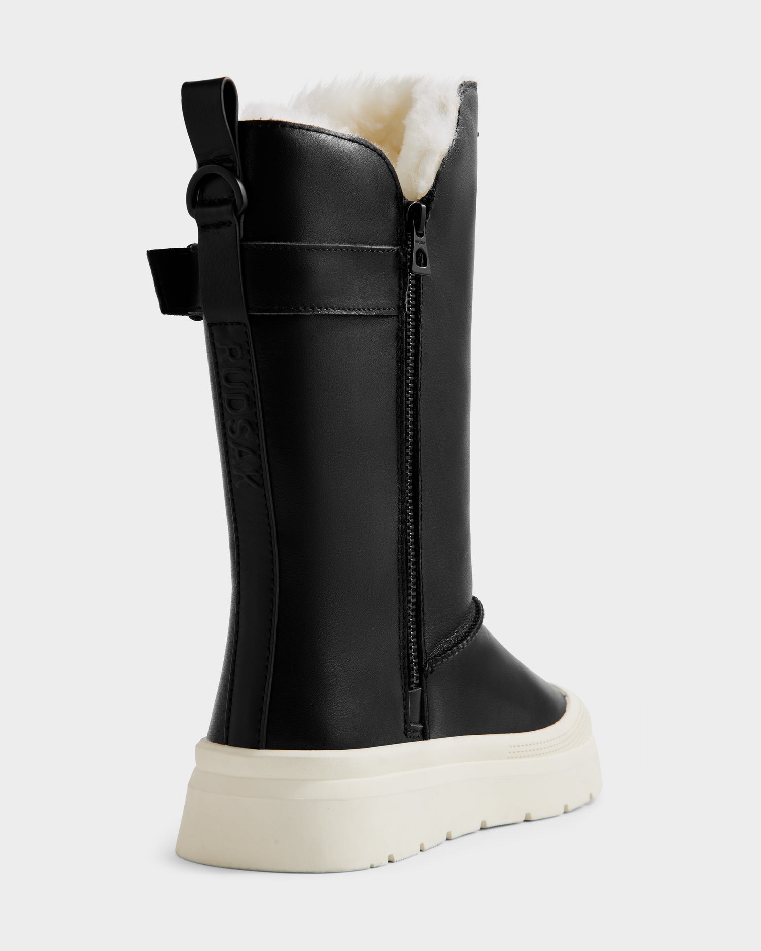 The KLAIR L - L225519 Black by Rudsak are women’s black mid-calf leather boots featuring a side zipper, white faux fur lining, pull tab, embossed branding, and a thick white sole for city-ready traction. Shown angled from the back and side.