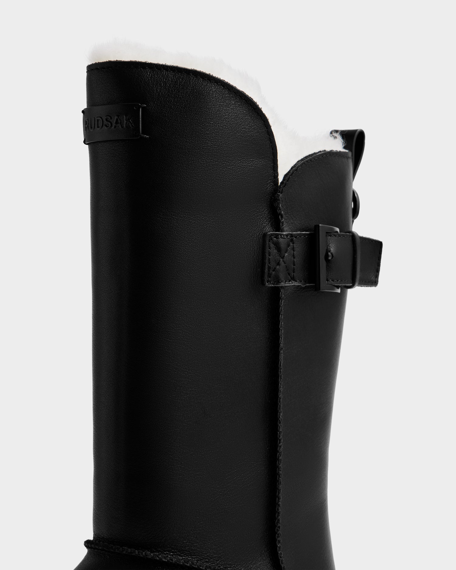 A close-up of the Rudsak KLAIR L - L225519 black women’s leather boot with white fur lining, a side buckle, pull tab, and city-ready traction. "RUDSAK" is embossed near the top.