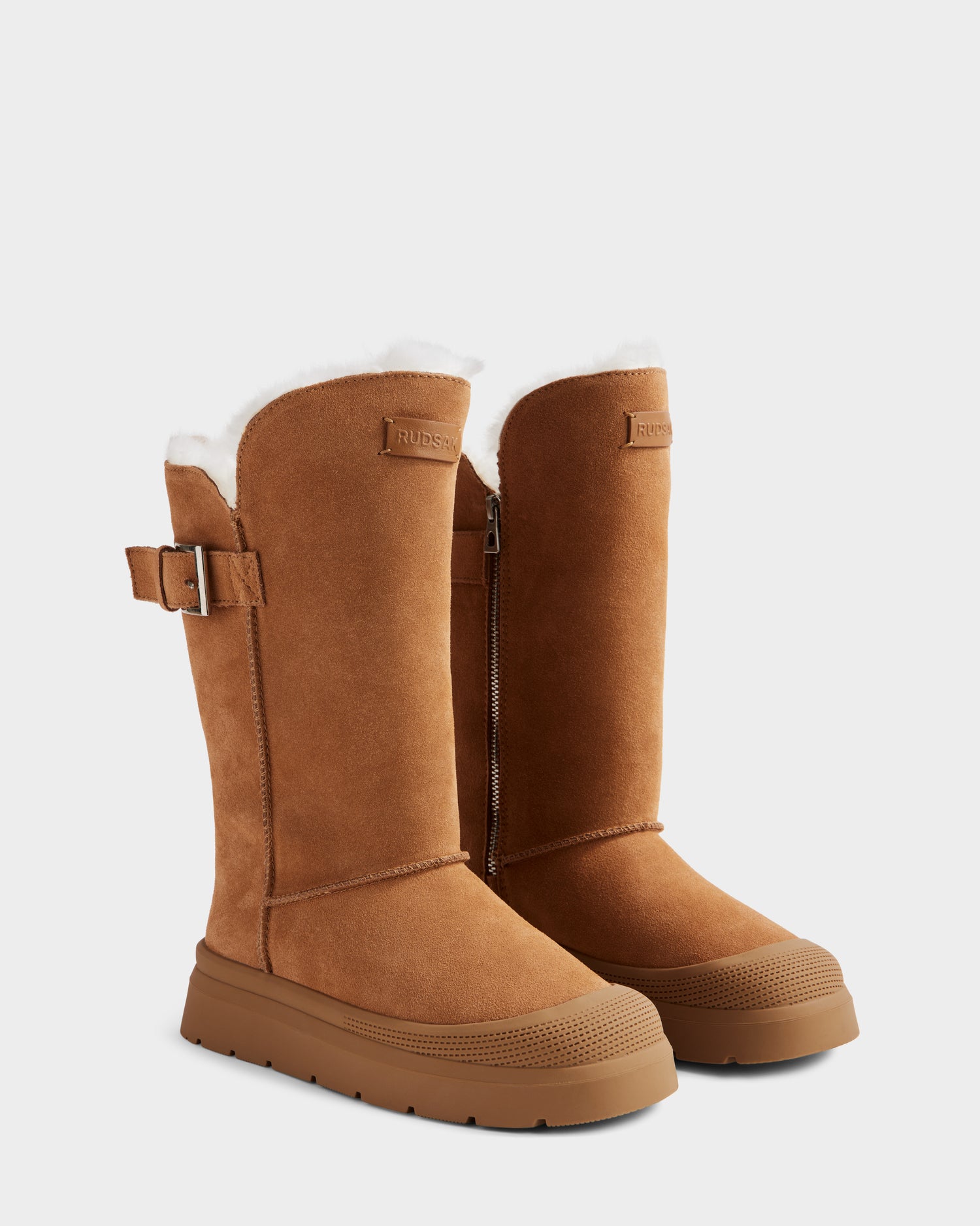 The Rudsak KLAIR - L225027 Tan women’s mid boots feature tall tan suede uppers, rubber soles, white wool lining, side zippers, and decorative buckles near the top, shown against a plain light background.