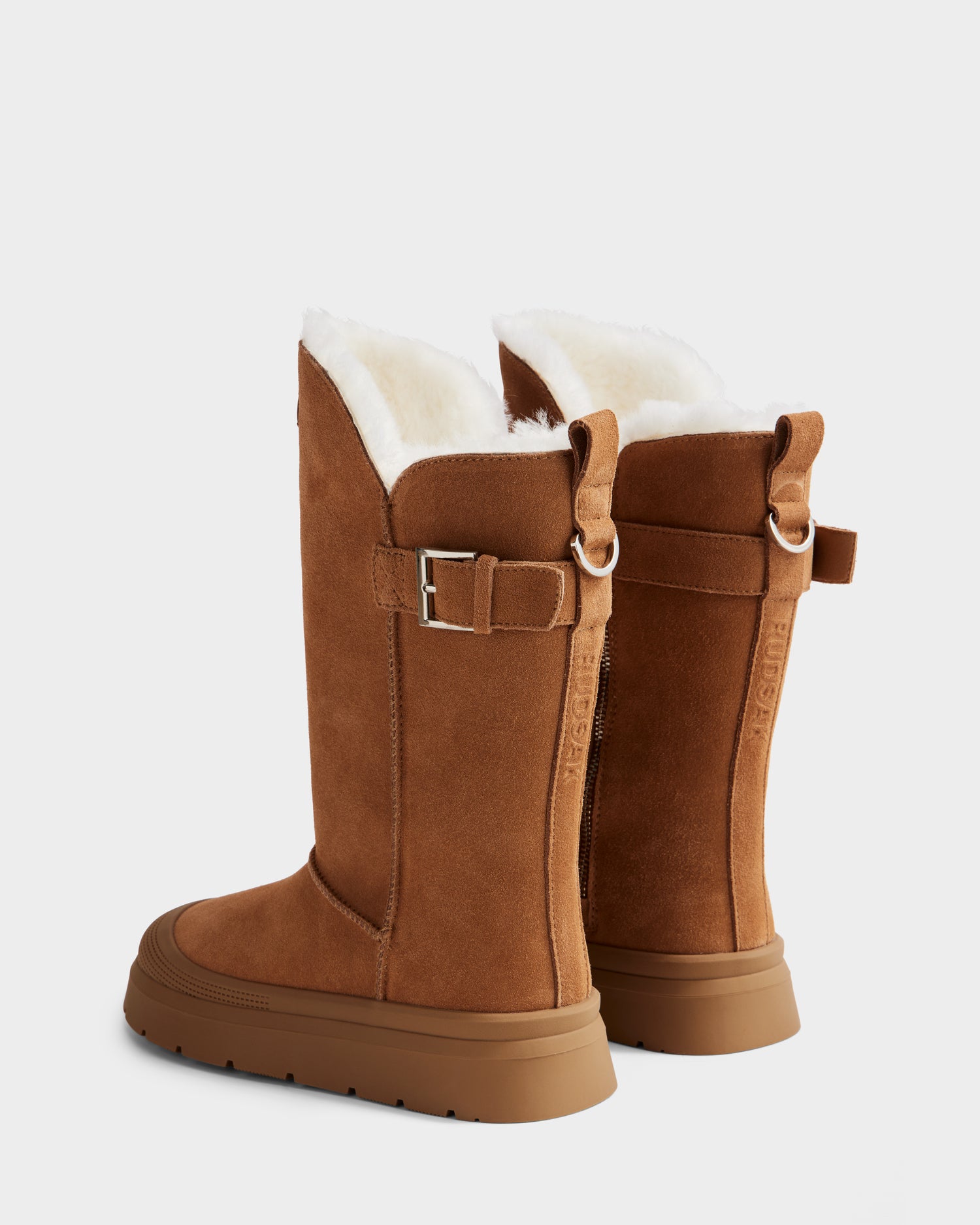 The Rudsak KLAIR - L225027 Tan boots feature tall tan suede, white faux fur lining, buckled straps, and ridged brown soles for stylish comfort on chilly days. Wool lined, they are perfect winter boots shown against a light background.