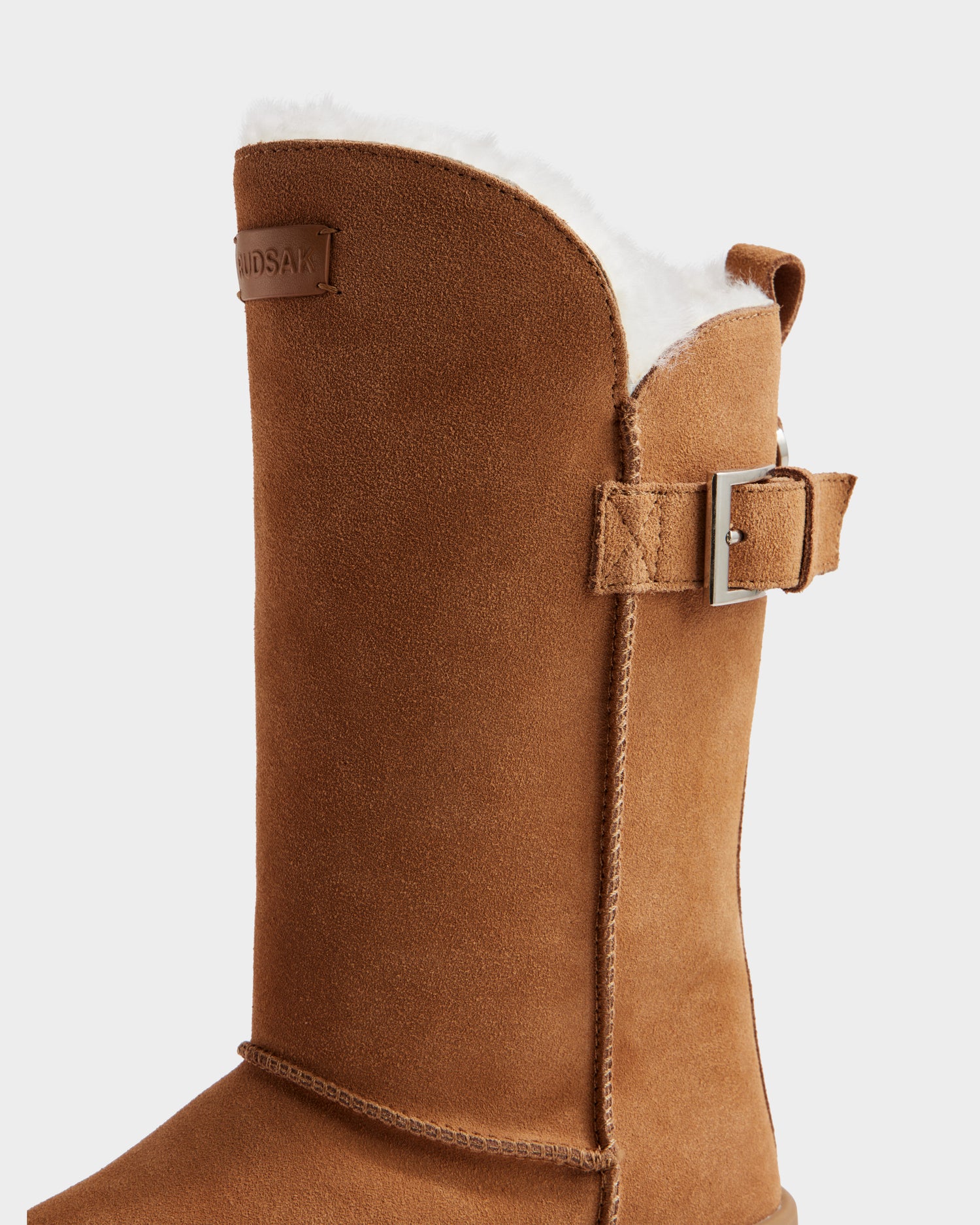 Close-up of the Rudsak KLAIR - L225027 Tan women’s mid boot in suede, with white fleece lining, decorative side buckle, and brand label near the top—a stylish choice for colder days.