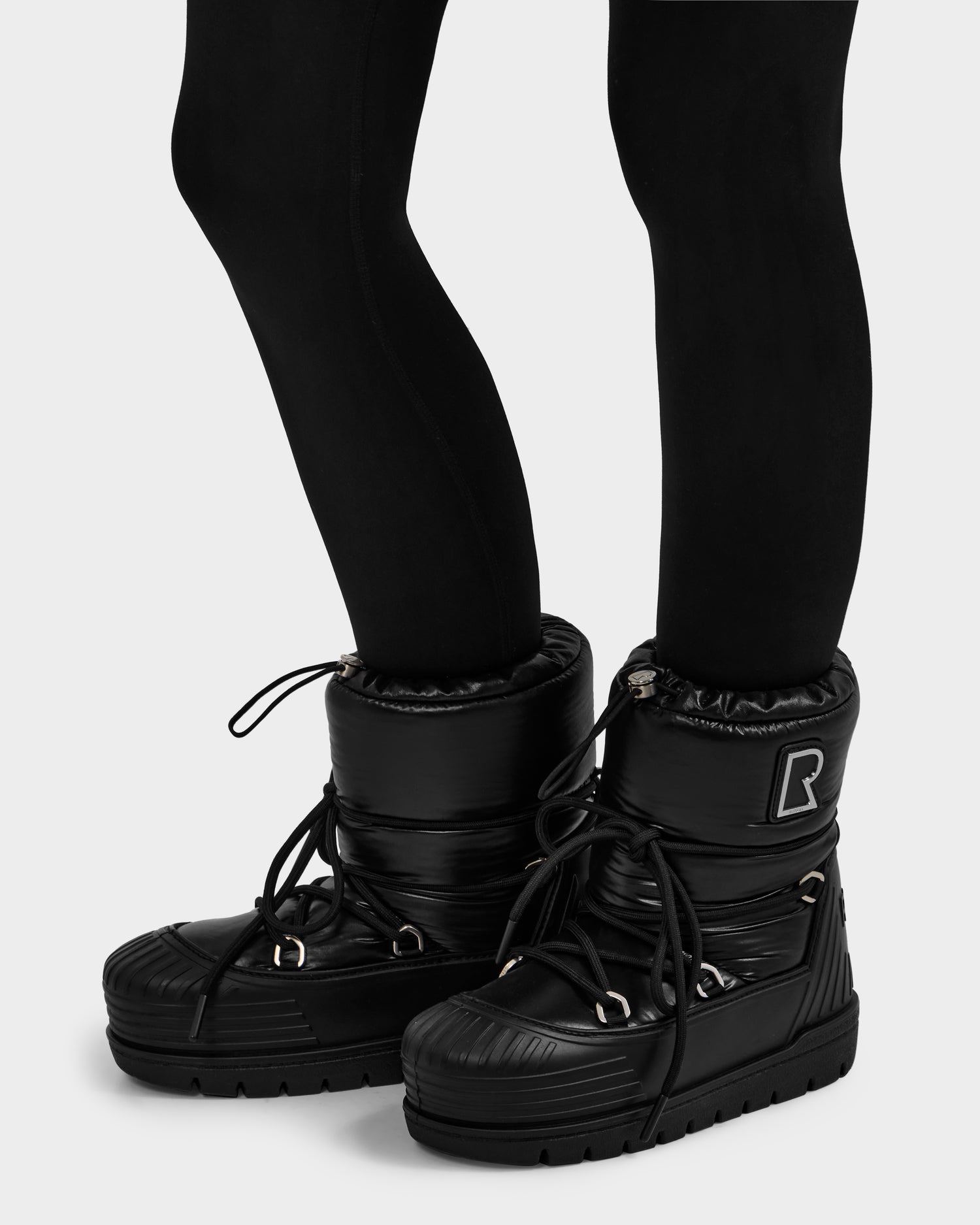 A person wearing black leggings and Rudsak KIKA - L224506 Black women's winter boots featuring thick laces, a rubber sole, and a stylized R logo—perfect for winter adventures.