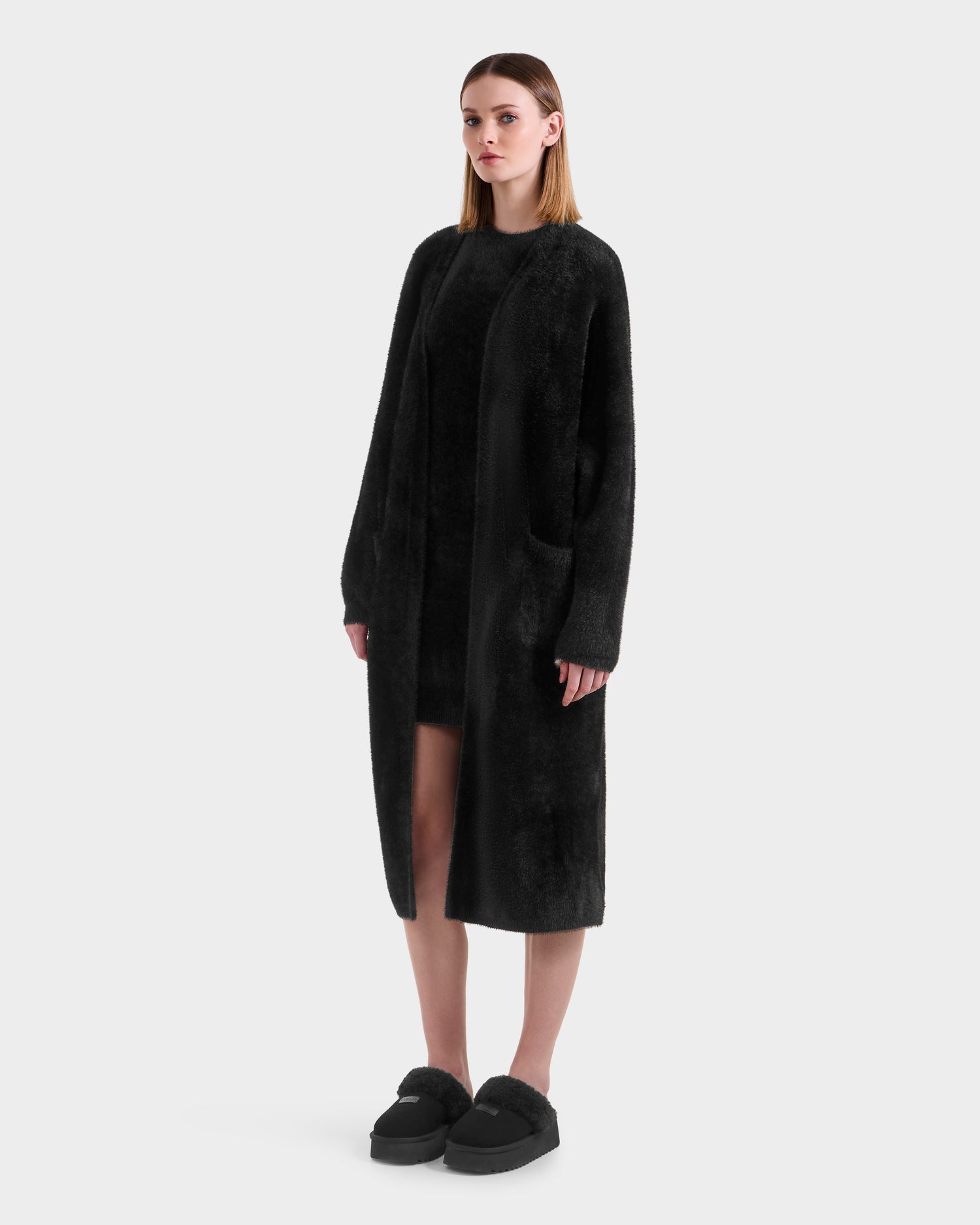 A woman with straight, shoulder-length hair stands facing forward in a neutral pose, wearing the Rudsak LACEY - 8525694 Black women’s fuzzy cardigan over a black dress and slippers against a plain white background.