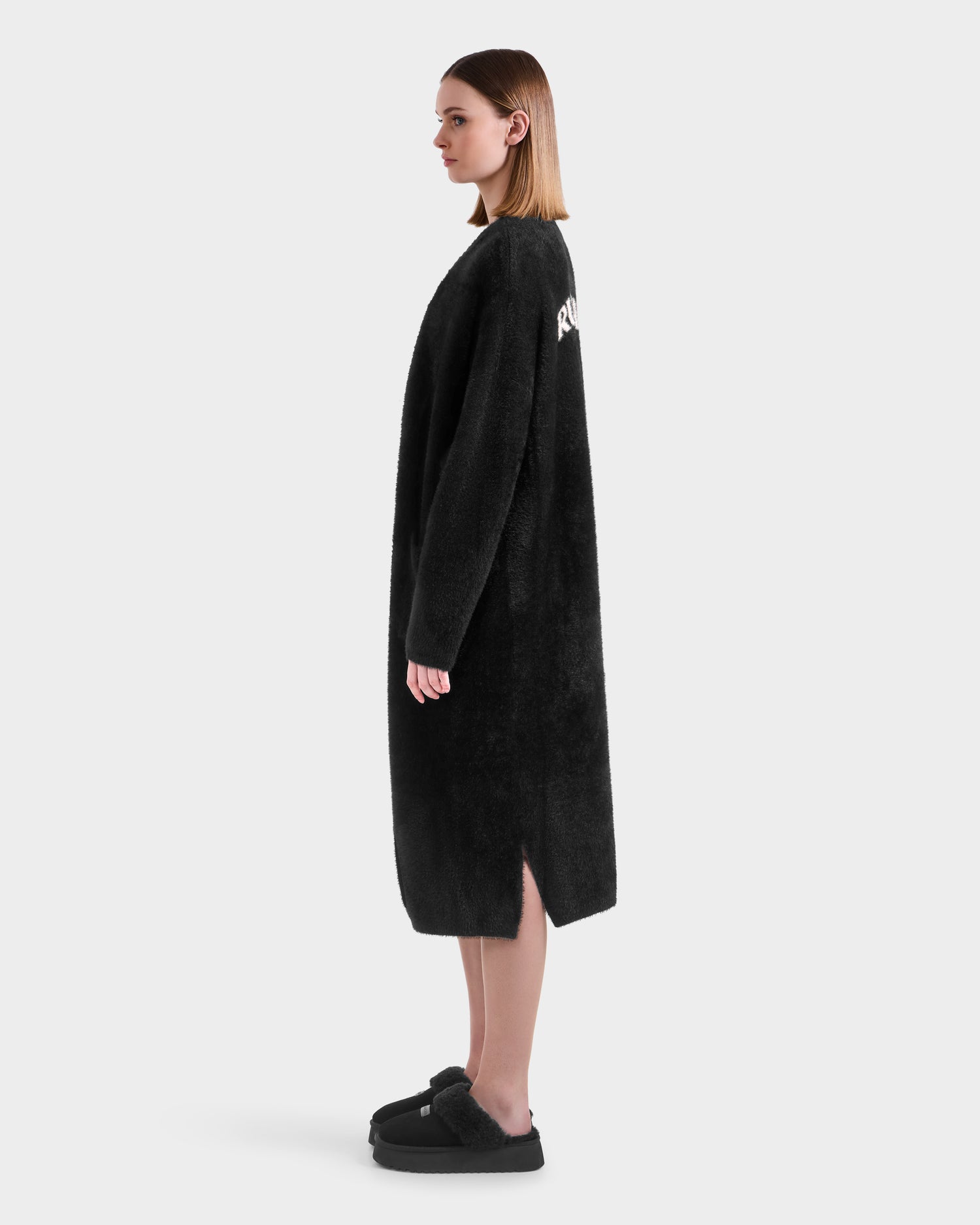 A woman stands in profile wearing the Rudsak LACEY - 8525694 Black fuzzy cardigan with side slits and matching black slip-on shoes, against a plain light background.