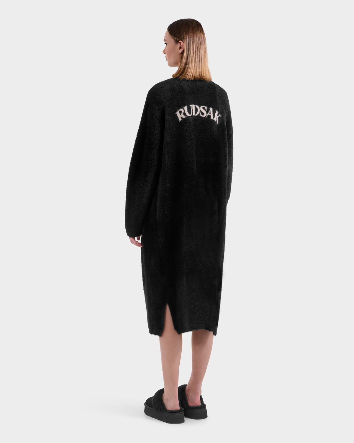 A person faces away, wearing the Rudsak LACEY - 8525694 black women’s fuzzy cardigan coat with “RUDSAK” in white on the back, side slits, and black platform shoes. The background is plain white.