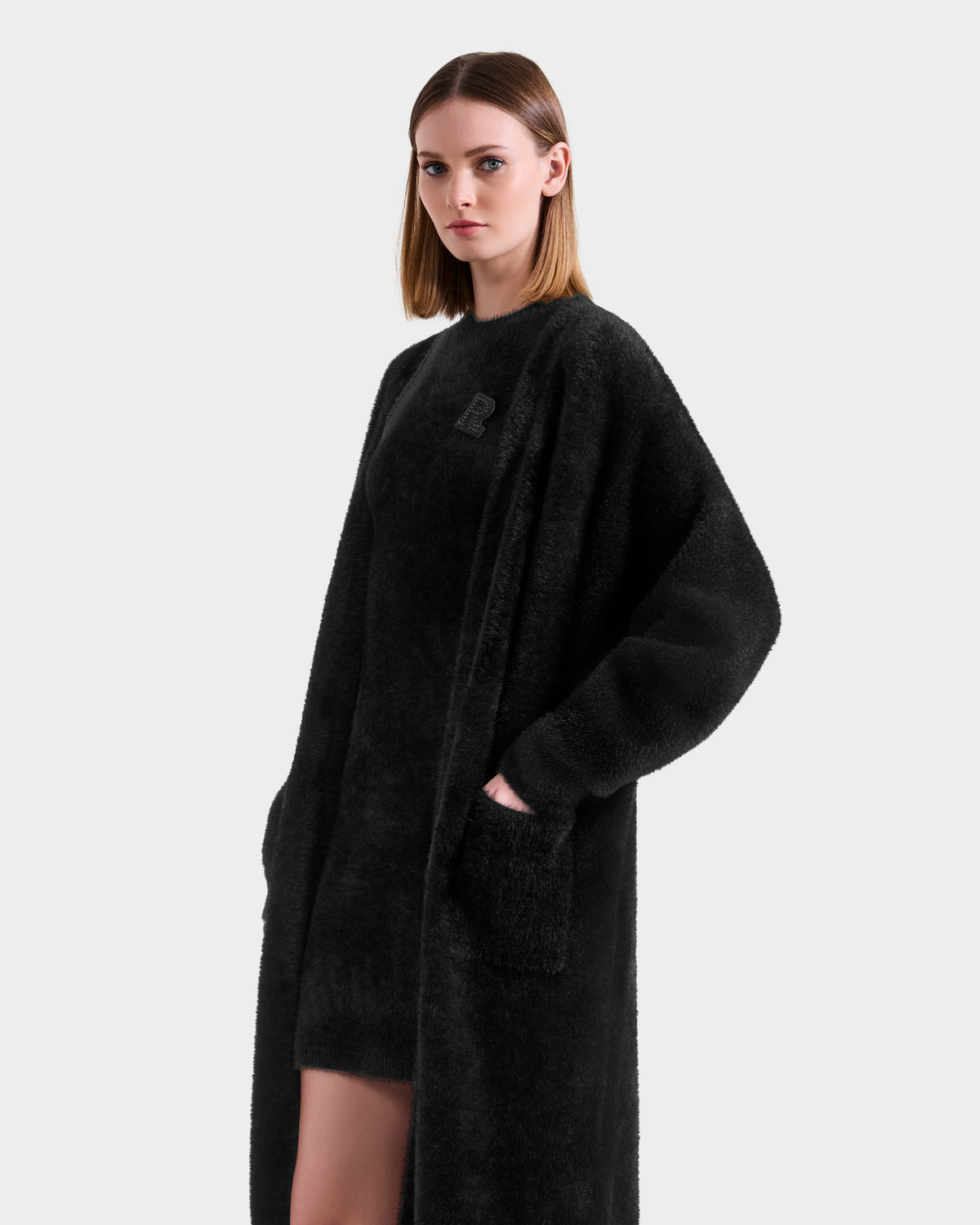 A woman with straight, shoulder-length hair wears the Rudsak LACEY - 8525694 Black long, textured coat over a cozy knit sweater and matching black outfit. She stands against a plain light gray background, looking slightly to the side.