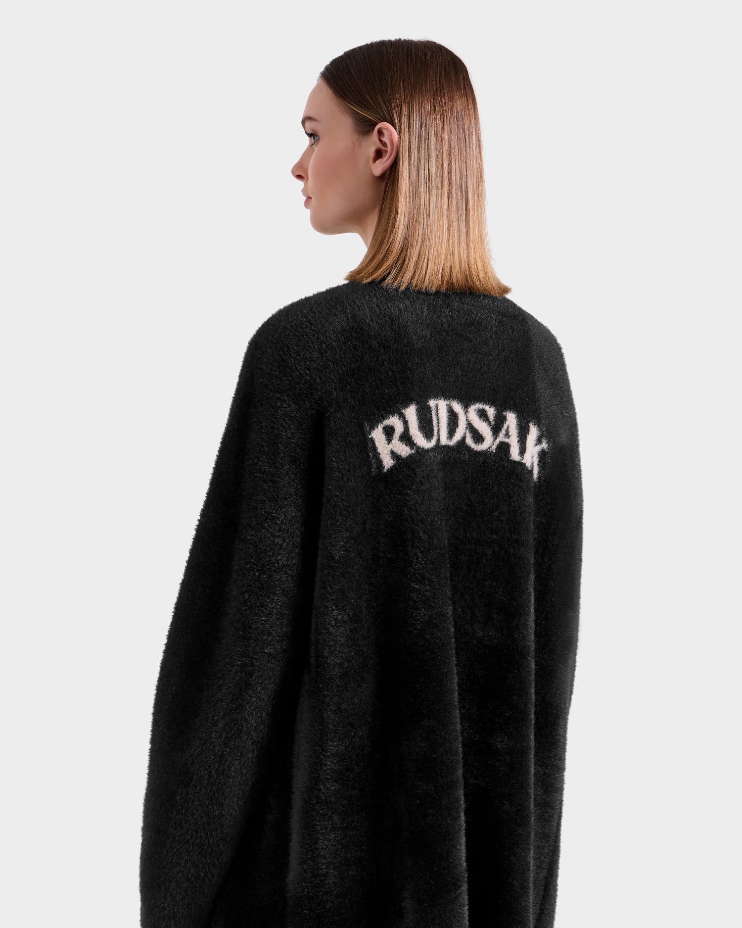 A woman with straight, shoulder-length hair models the LACEY - 8525694 Black fuzzy cardigan by Rudsak, featuring the brand’s name in white on the back, against a plain light gray background.