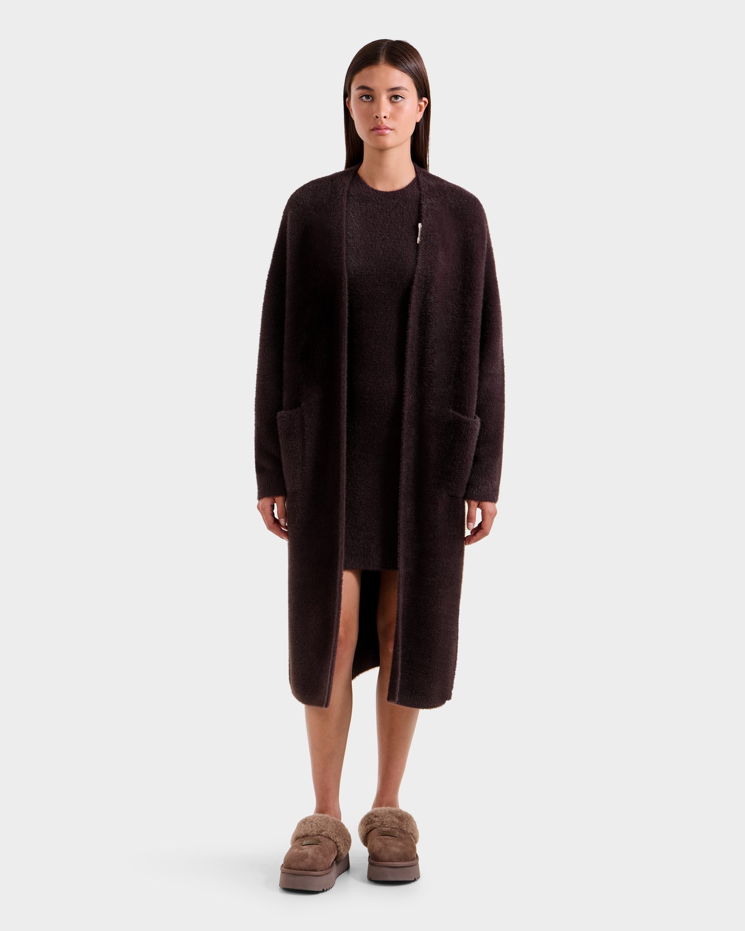 A woman with straight brown hair and a neutral expression models the Rudsak LACEY - 8525694 Espresso cardigan sweater, layered over a matching dress and paired with fuzzy brown slippers, against a plain white background.