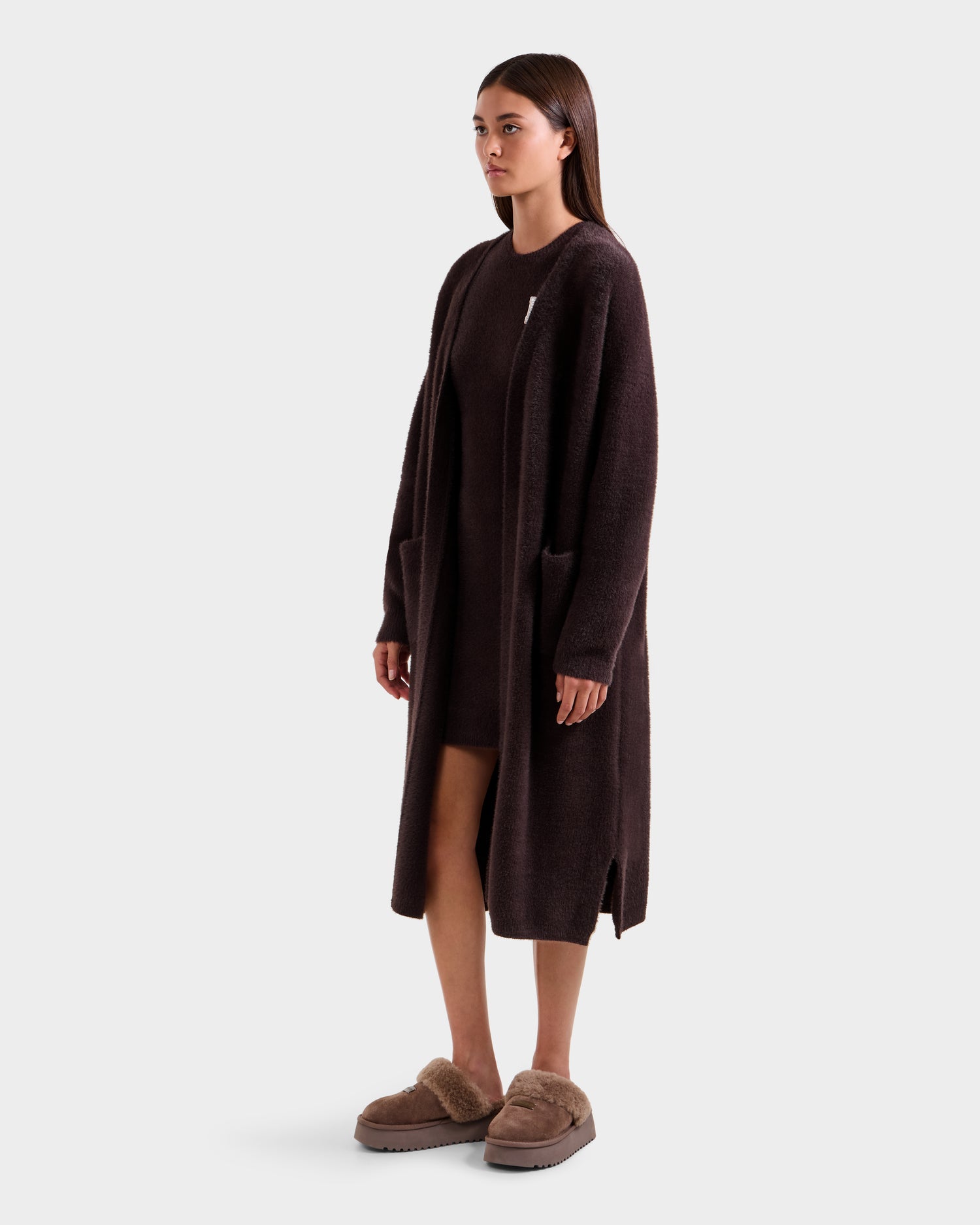 A woman stands against a plain background wearing the Rudsak LACEY - 8525694 Espresso fuzzy cardigan, layered over a matching dress with tan fuzzy slippers. She looks ahead, arms relaxed, embracing cozy layering for comfort.