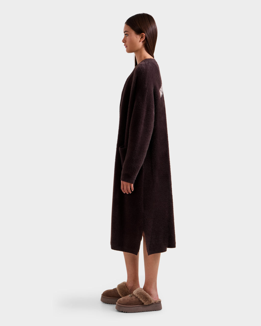 A woman stands in profile against a plain light gray background, wearing the Rudsak LACEY - 8525694 Espresso long cardigan sweater with pockets and cozy brown fur-trimmed slippers, perfect for layering.