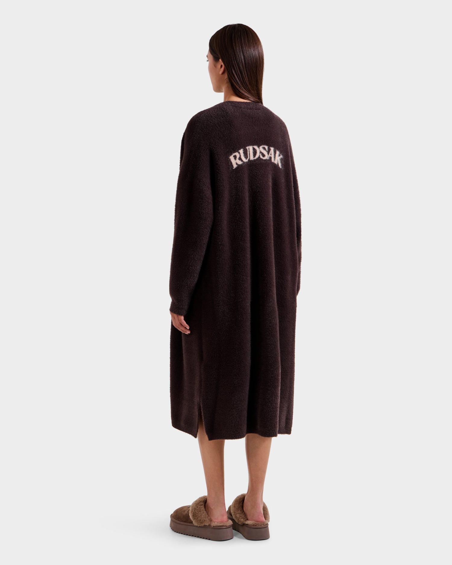 A person stands facing away, wearing the Rudsak LACEY - 8525694 espresso cardigan with RUDSAK on the back and matching slippers. The cozy piece stands out against a plain white background.