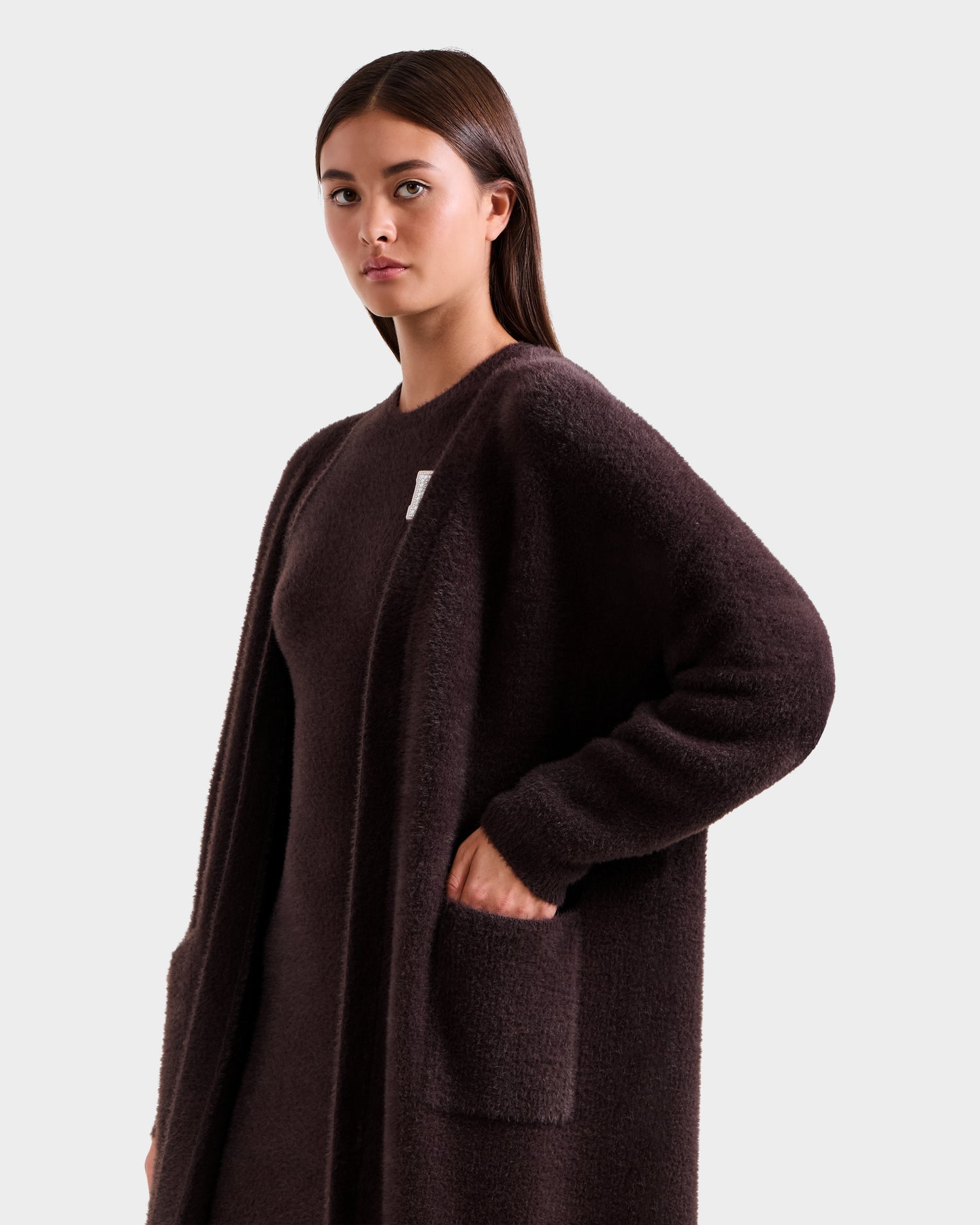 A woman with straight brown hair wears the Rudsak LACEY - 8525694 Espresso cozy long cardigan sweater with large front pockets over a matching top, standing against a plain light grey background.