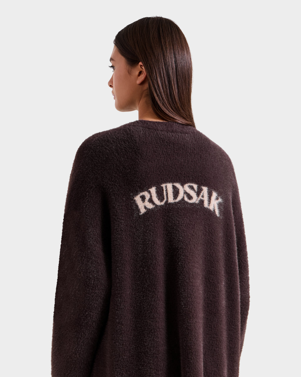 A woman with straight brown hair wears the Rudsak LACEY - 8525694 Espresso dark fuzzy coat, perfect for cozy layering, featuring "RUDSAK" in white on the back, as she stands facing left against a plain light background.